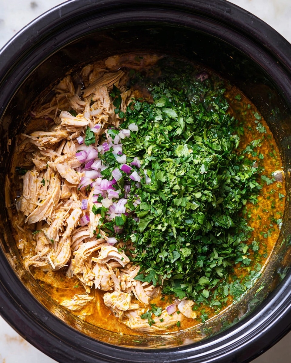 In a black cooking pot, there is one main layer of light brown shredded cooked chicken mixed with a thick orange-brown sauce at the bottom. On top of the chicken and sauce, a thick pile of deep green chopped fresh herbs, likely cilantro, covers about half the surface on the left side. Scattered on the shredded chicken and mixed with the herbs are small pieces of finely chopped purple onion. The pot is placed on a white marbled surface. photo taken with an iphone --ar 4:5 --v 7