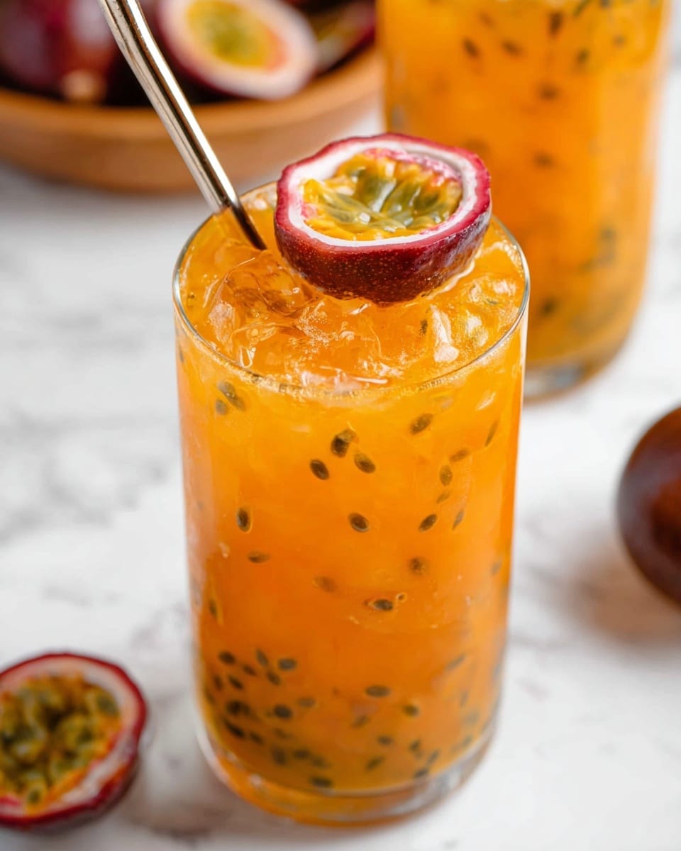 A tall clear glass filled with a bright orange drink that has black passion fruit seeds scattered throughout. The drink has ice cubes, giving a textured look near the top. On top of the ice sits a half piece of passion fruit, showing its yellow, pulpy inside with black seeds and a red edge. A metal straw is placed diagonally inside the glass, resting near the top left. The glass sits on a white marbled surface, with blurred matching drinks and a whole passion fruit nearby. Photo taken with an iphone --ar 4:5 --v 7