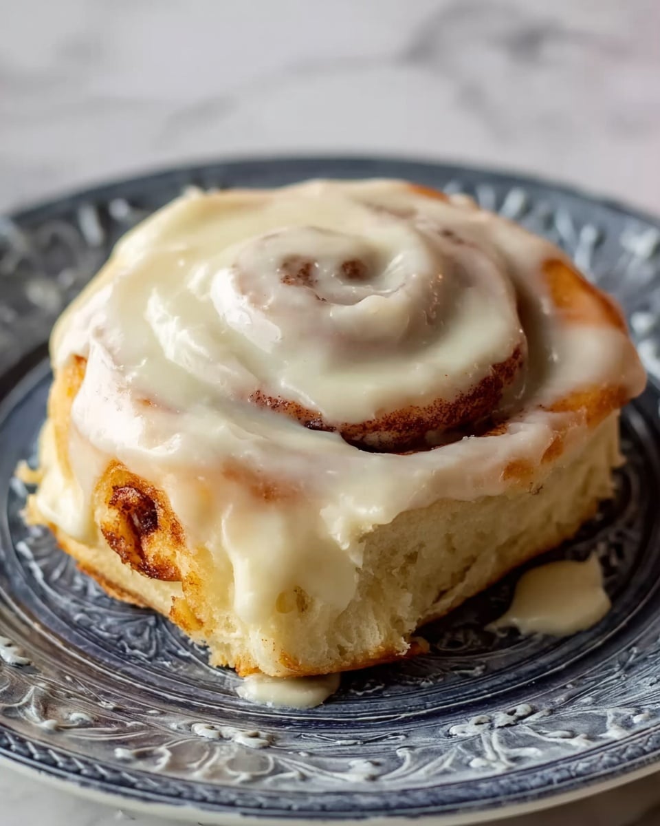A single cinnamon roll sits in the center of a white plate with decorative raised patterns, placed on a white marbled surface. The cinnamon roll has three visible layers: the bottom layer is a thick, soft dough with a light brown baked edge; the middle layer shows the spiral swirl of cinnamon filling, dark brown in color; the top layer is a smooth, creamy off-white icing generously spread, slightly dripping over the sides. The icing covers most of the roll's surface but reveals the spiral below. The lighting highlights the soft texture of the dough and the shine of the icing. Photo taken with an iphone --ar 4:5 --v 7