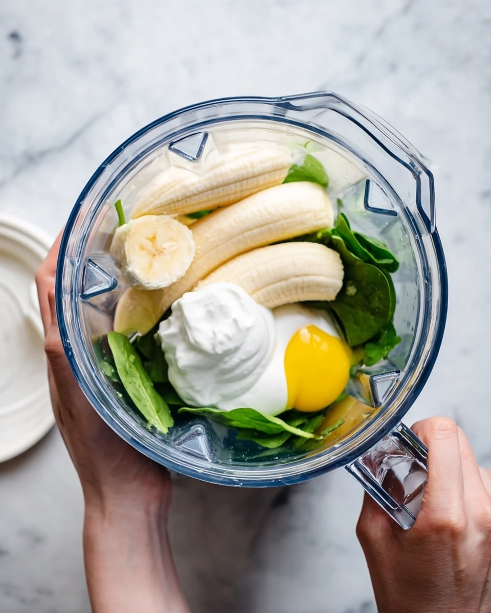 A clear blender jar held by a woman's hands contains several ingredients layered together: at the bottom, fresh green spinach leaves create a leafy base, above that two peeled whole bananas lay side by side with a smooth, creamy yellow-white texture, next to a single bright yellow egg yolk with clear egg whites around it, and on top, a dollop of thick white yogurt sits. The lid of the blender lies on the white marbled surface nearby. The photo taken with an iphone --ar 4:5 --v 7