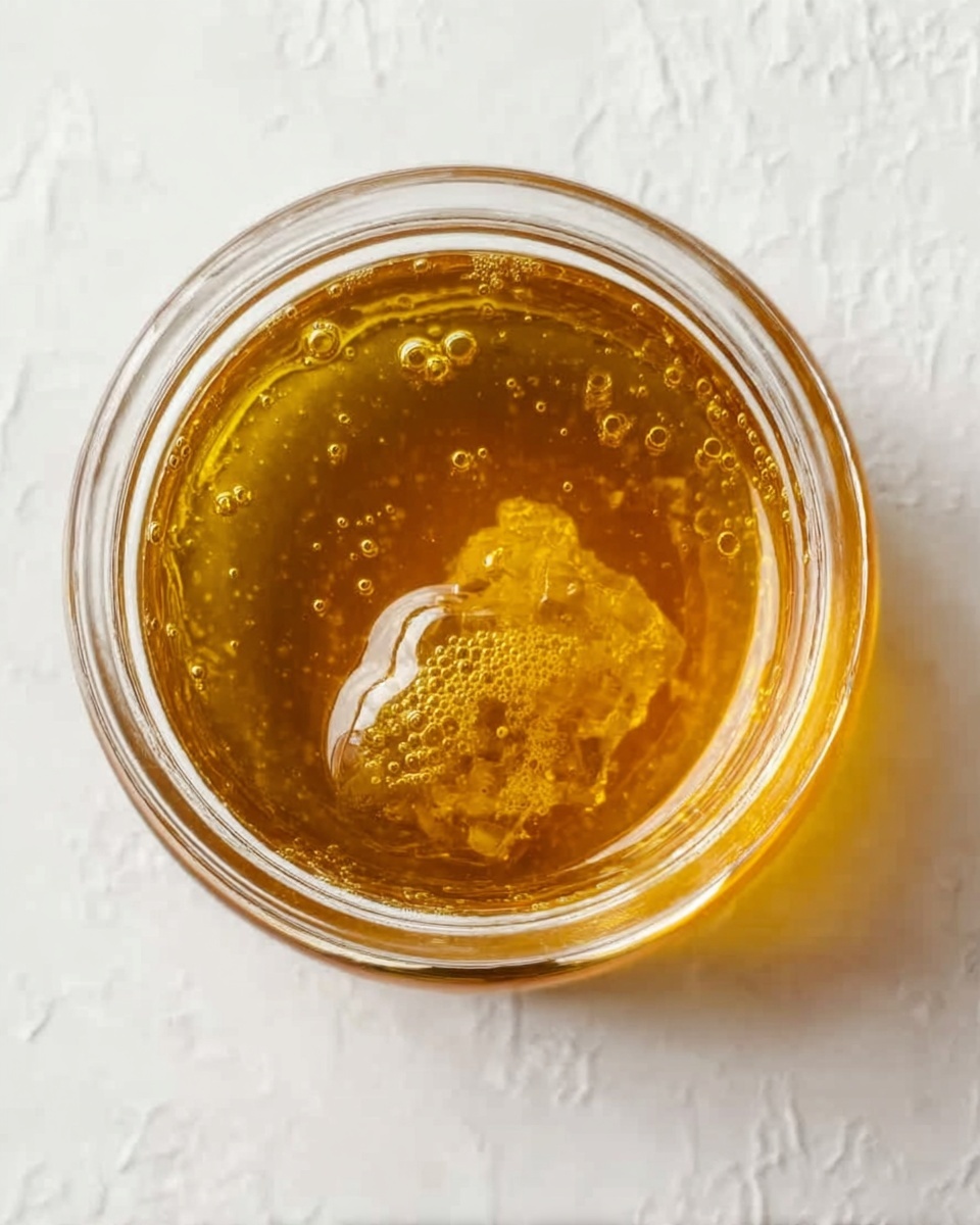 A close-up top view of a small clear glass jar filled with golden honey. The honey is thick and shiny with small air bubbles trapped inside, and a piece of honeycomb floats near the center. The jar sits against a white marbled textured surface with soft natural light highlighting the smooth, sticky texture of the honey. Photo taken with an iphone --ar 4:5 --v 7