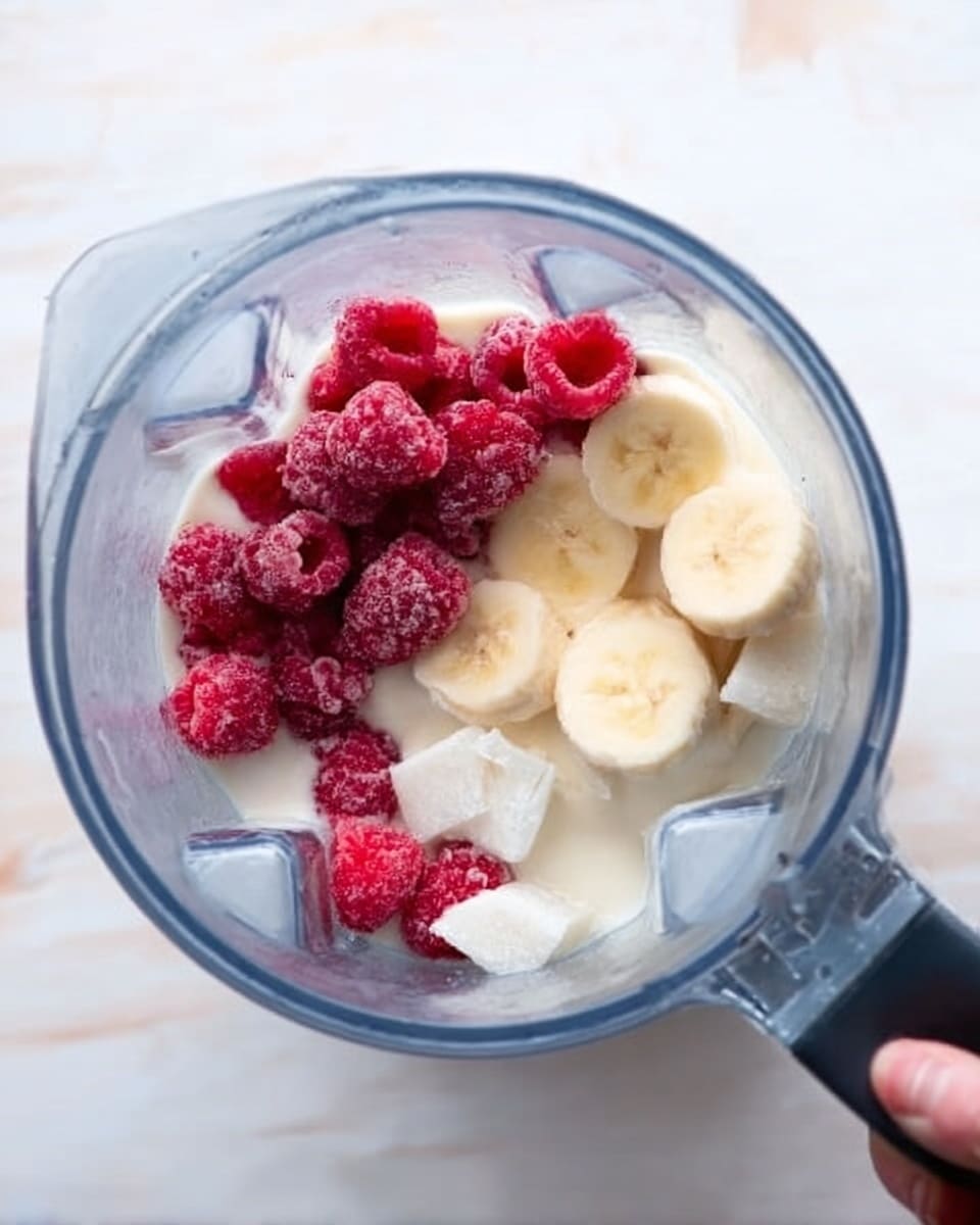 Inside a clear blender jar, there are three main layers of ingredients visible from top to bottom. The top layer has bright red frozen raspberries grouped together on the left side, showing a frosty texture. Below and to the right of the raspberries, there are round banana slices with a pale yellow color that look soft and fresh. The banana slices sit on a creamy white layer of yogurt or milk that fills most of the container base. Small chunks of white frozen cubes, likely coconut or ice, are scattered around the other ingredients. The blender jar is placed on a white marbled surface. A woman’s hand is holding the blender. Photo taken with an iphone --ar 4:5 --v 7