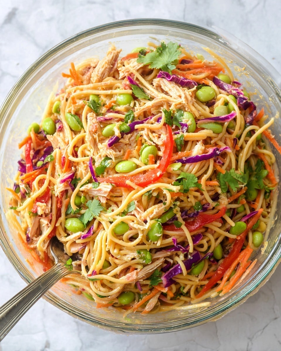 The dish is a colorful noodle salad served in a clear glass bowl, showing a mix of light yellow noodles as the base layer, with bright green edamame beans and shredded orange carrots scattered throughout. Thin strips of red bell pepper and purple cabbage add vibrant color, mixed evenly with the noodles. Small pieces of shredded light brown chicken are spread within the salad. Fresh green cilantro leaves are sprinkled on top, adding a touch of freshness. A metal spoon is partially visible on the left side of the bowl. The background features a white marbled texture. Photo taken with an iphone --ar 4:5 --v 7