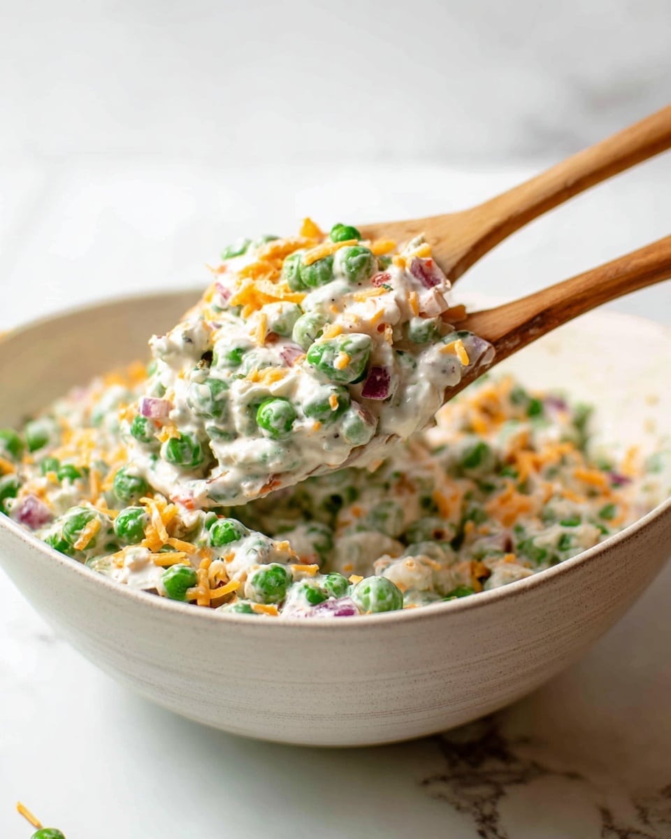 A large white bowl filled with a creamy salad that has visible green peas, shredded orange cheese, and small pieces of red onion mixed in a white dressing. Two wooden spoons lift a portion of the salad above the bowl, showing the thick texture and colorful mix of ingredients against a white marbled surface. photo taken with an iphone --ar 4:5 --v 7
