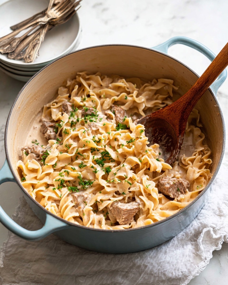 A close-up view of a large light blue pot filled with creamy pasta and chunks of meat. The pasta is egg noodles, light yellow with a smooth, soft texture, mixed evenly throughout the creamy sauce that is light beige with a slightly glossy surface. Scattered green parsley pieces add a touch of color on top. There are several pieces of brown, tender meat nestled within the noodles. A wooden spoon with a natural brown color rests inside the pot, partially covered in sauce. The pot sits on a white marbled surface with a white cloth underneath. Nearby are stacked plain white bowls and two slightly tarnished silver forks. photo taken with an iphone --ar 4:5 --v 7