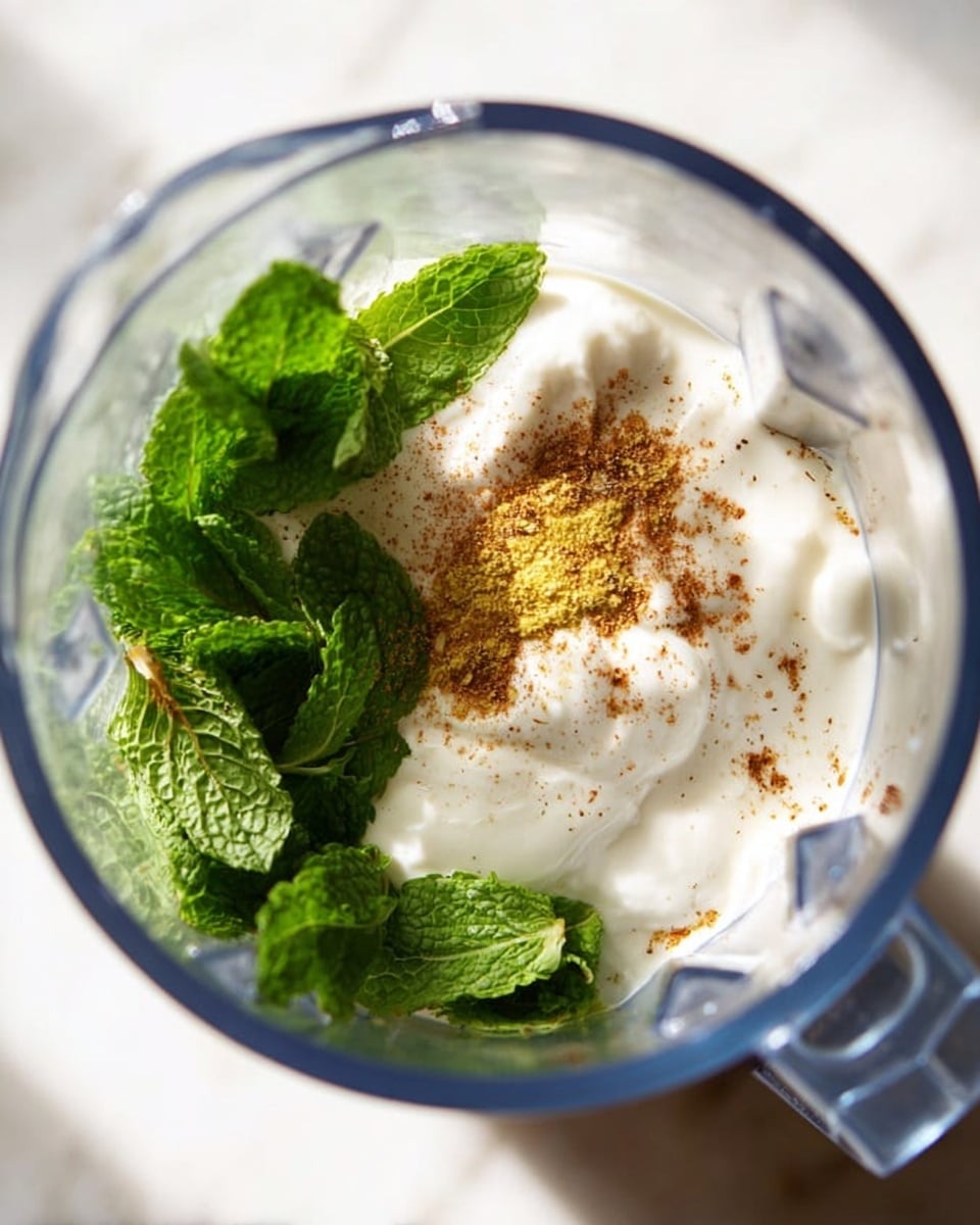The image shows the inside of a blender with three layers visible: at the bottom, fresh green mint leaves with detailed texture; above the leaves is a thick, creamy white yogurt layer with a smooth texture; on top of the yogurt, there is a sprinkle of brown and yellow spices lightly scattered in the center. The blender itself has a transparent body and a handle that appears blue. The photo has a soft, bright lighting and the whole scene is on a white marbled surface. Photo taken with an iphone --ar 4:5 --v 7