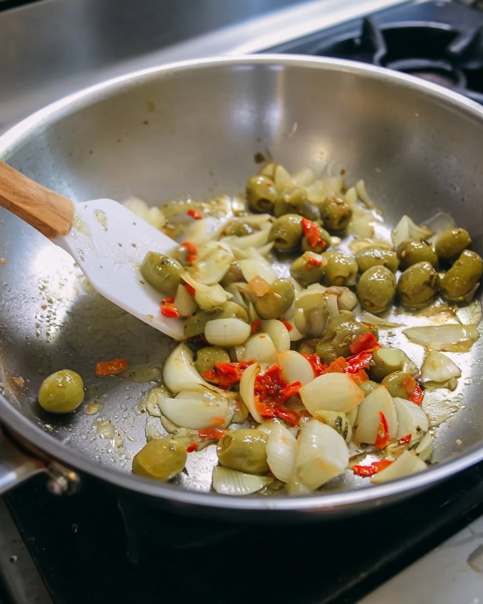 A silver frying pan showing small pieces of green olives, thin white slices of garlic, and small bits of red chili pepper being cooked. A white spatula with a wooden handle is scooping some of the ingredients from the left side of the pan. The pan is on a stove with a black burner visible in the background. The mixture looks shiny with a light coating of oil. The pan has a smooth and slightly reflective surface. photo taken with an iphone --ar 4:5 --v 7