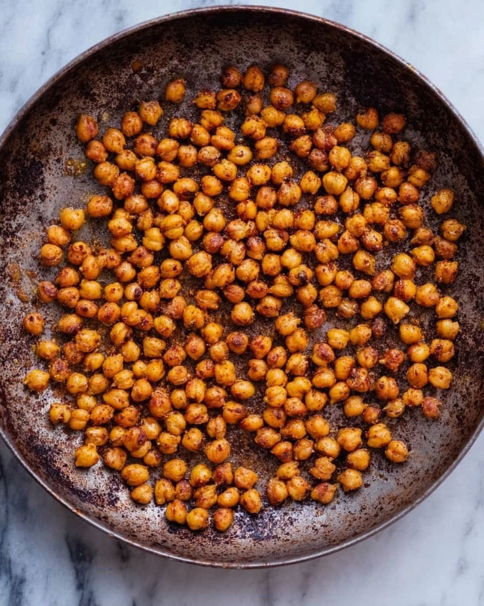 A round dark metal pan holds a single layer of small, golden-brown roasted chickpeas spread evenly across the surface. The chickpeas have a slightly rough texture with some darker spots, indicating a crispy outer layer. The pan shows signs of use, with patches of rust and discoloration adding to the rustic look. The background is a white marbled surface. photo taken with an iphone --ar 4:5 --v 7