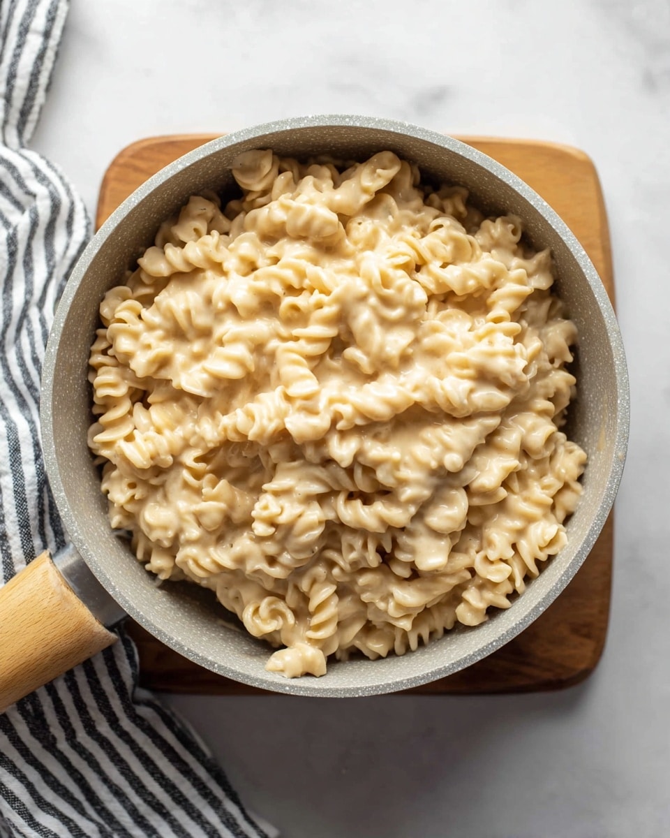A pot full of curly pasta covered in a creamy, light beige cheese sauce. The pasta is tightly packed with the sauce evenly coating every piece, giving it a smooth and rich look. The pot is light gray with a wooden handle, sitting on a small, square wooden board. The background surface has a white marbled texture, and a striped cloth is partially visible at the top corners. photo taken with an iphone --ar 4:5 --v 7