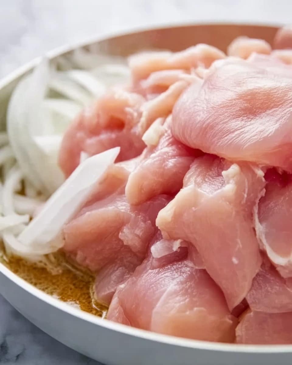A close-up view of a white pan filled with raw, light pink chicken pieces piled in the center, showing soft and smooth textures with some layers overlapping on top of each other. On the side, there are some thin slices of white onion resting against the chicken. The background features a white marbled texture. Photo taken with an iphone --ar 4:5 --v 7