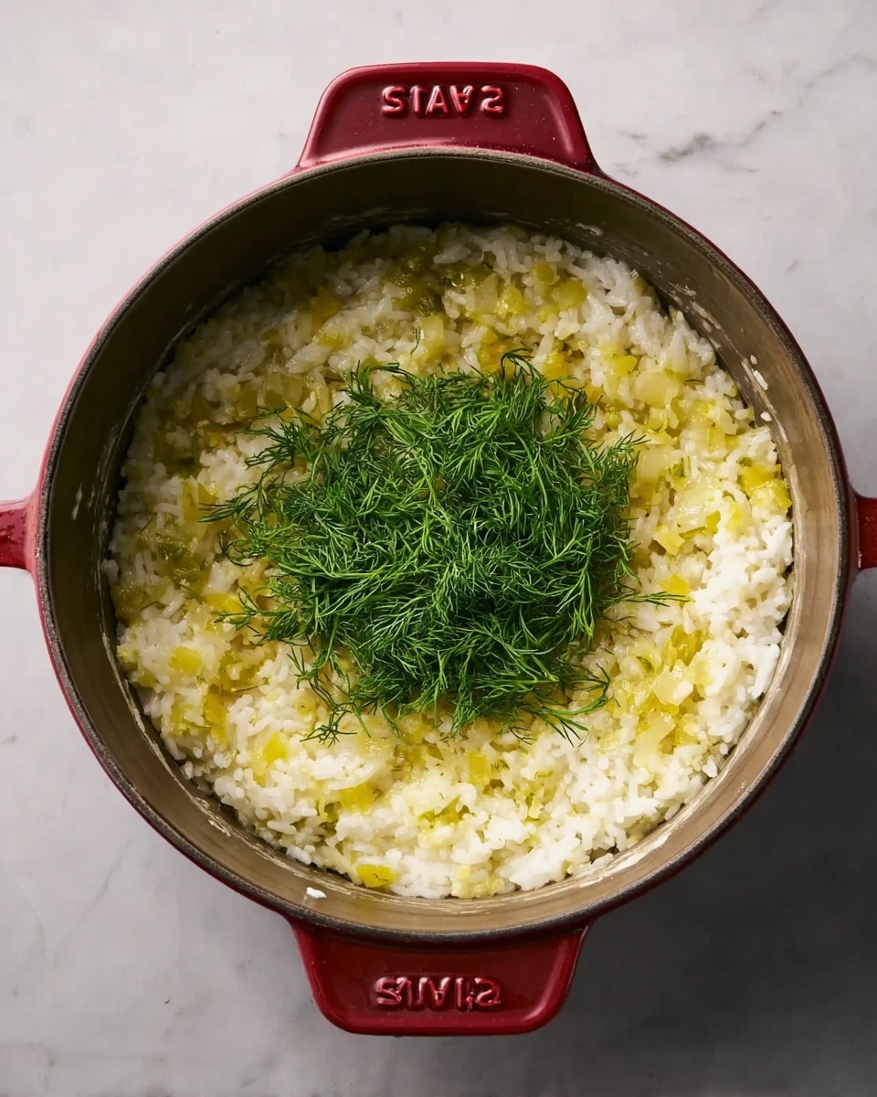 A red Staub pot containing a creamy rice dish with small, diced yellow pieces mixed evenly throughout the white rice layer at the base. In the center of the pot, a fresh pile of bright green dill sits on top, contrasting with the creamy yellow and white mixture surrounding it. The pot handles have embossed red text on them. The pot is set on a white marbled surface. photo taken with an iphone --ar 4:5 --v 7