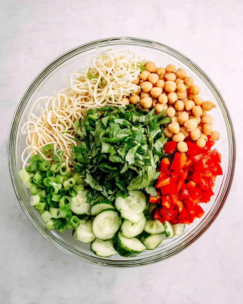 A clear glass bowl placed on a white marbled surface holds six distinct layers of ingredients arranged side by side. On the left is a small pile of thin, white spiraled noodles followed by a cluster of pale beige beans. Next to the beans is a big lump of fresh chopped green herbs with a leafy texture. To the right of the herbs is a pile of round, light tan chickpeas. Adjacent to the chickpeas is a section of bright red diced bell peppers scattered with small green chopped scallions. Completing the circle on the bottom right are light green cucumber slices, cut thick with dark green skin. The colors are fresh and vibrant, with clear separation between each ingredient. photo taken with an iphone --ar 4:5 --v 7