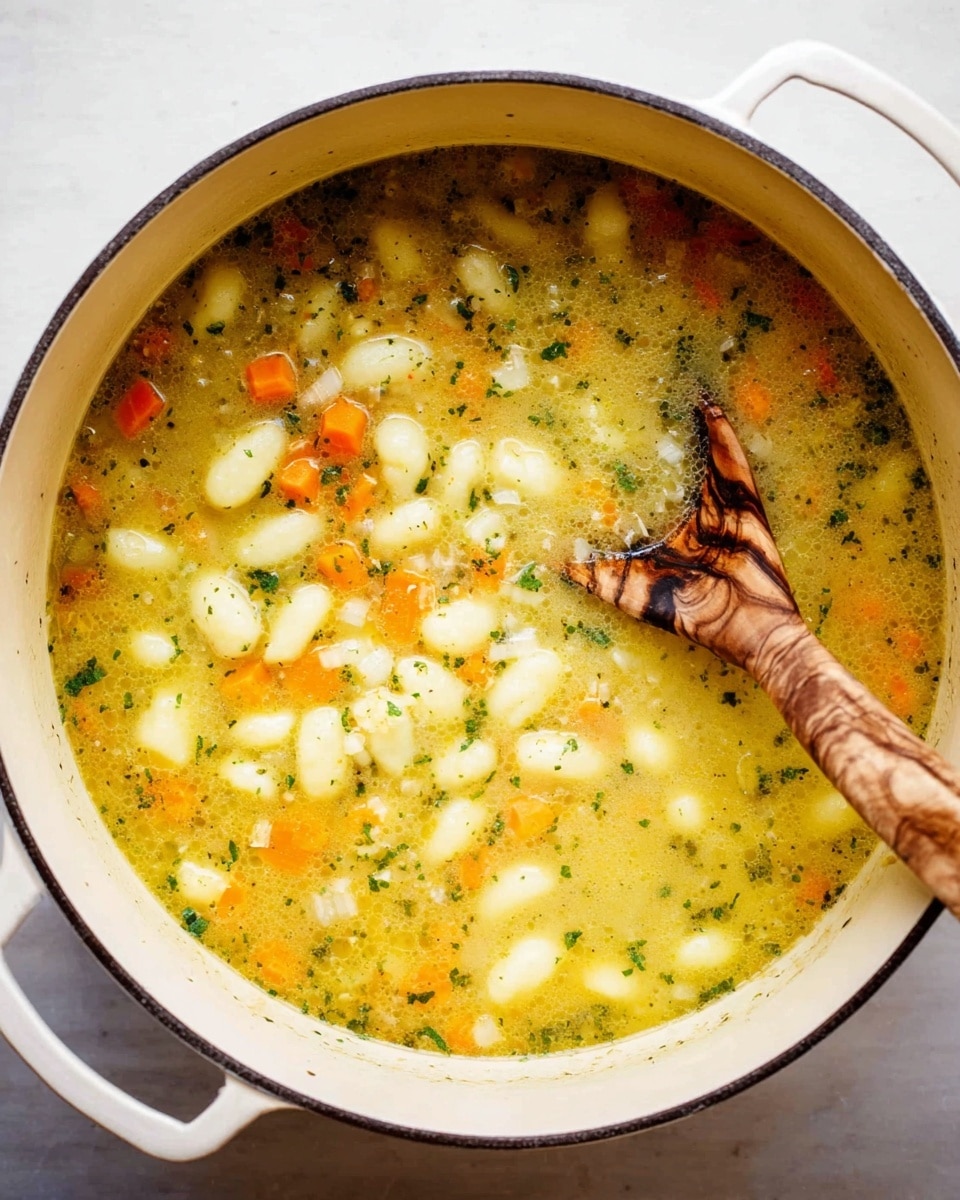 A white pot filled with a thick soup made of small gnocchi, diced orange carrots, and finely chopped onions scattered throughout a light yellow broth with visible green herbs, giving the soup a textured look. A wooden spoon with natural brown swirls rests inside the pot, partially submerged in the soup. The pot is placed on a white marbled surface. Photo taken with an iphone --ar 4:5 --v 7