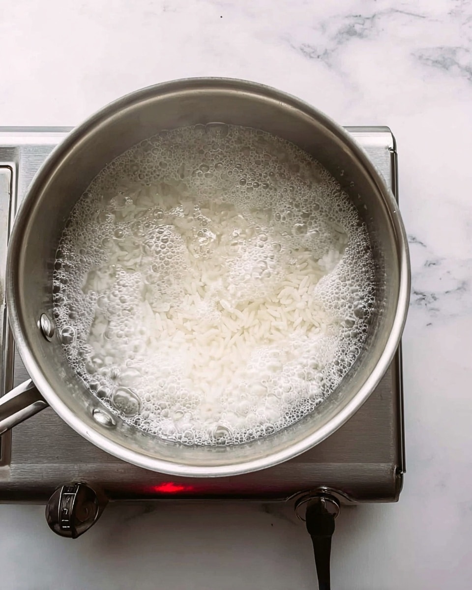 A metal pot is shown from above with white rice boiling inside clear water. The water has small bubbles and foam on the surface. The pot is placed on a silver electric stove on a white marbled surface with a small red light on the stove. photo taken with an iphone --ar 4:5 --v 7