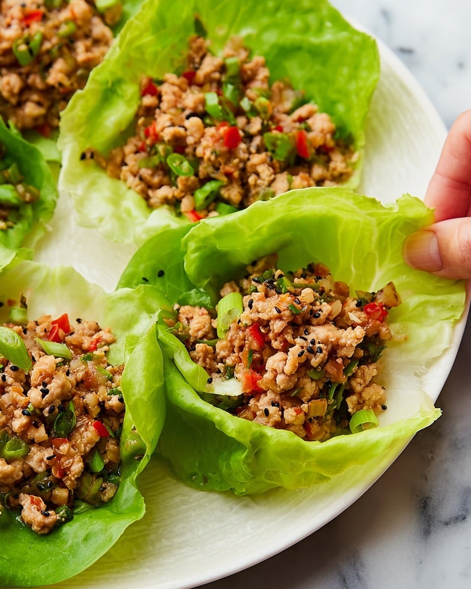 The image shows several lettuce cups arranged on a white plate with a white marbled surface underneath. Each lettuce cup is fresh and light green with a slightly crinkled texture, forming a natural bowl shape. Inside each cup, there is a filling made of finely chopped cooked meat mixed with small pieces of red pepper, green onion slices, and black sesame seeds, adding varied texture and color to the dish. The filling looks moist and slightly crumbly, contrasting with the crisp lettuce leaves. In the foreground, a woman's hand is touching one of the lettuce cups, making the scene more inviting. The overall look is fresh and colorful with a neat and appetizing arrangement photo taken with an iphone --ar 4:5 --v 7