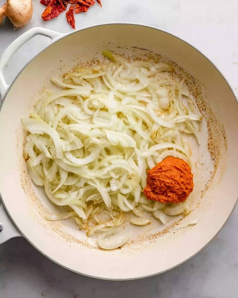 The image shows a white pan on a white marbled surface. Inside the pan, there are thin white onion slices spread out in a soft layer, slightly cooked. At the bottom right side of the onions, there is a small round ball of bright reddish-orange paste resting on the pan’s surface. The pan has light brown residue marks around the sides, indicating some cooking done. The background includes blurred small pieces of red and beige foods slightly visible at the top of the frame. Photo taken with an iphone --ar 4:5 --v 7