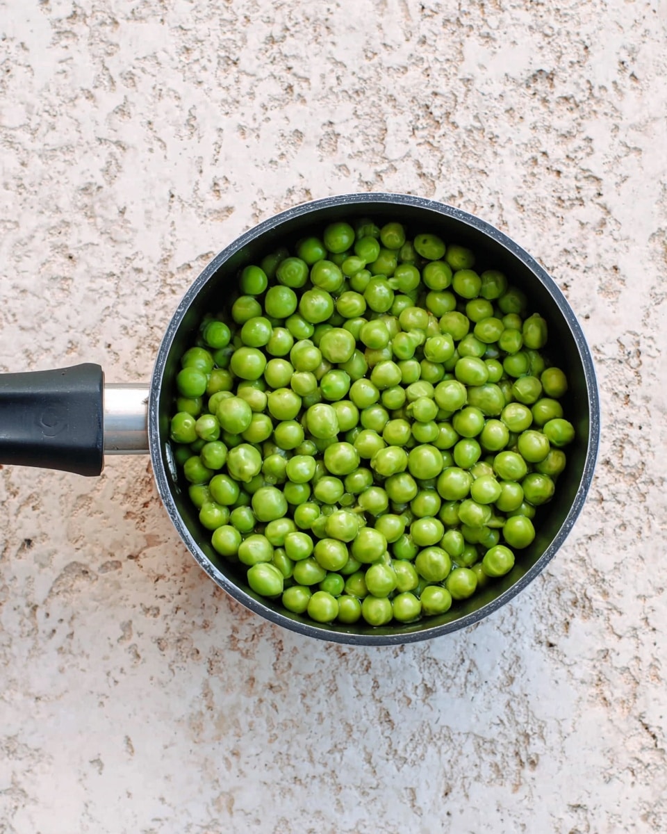 A small black pot filled with a single layer of fresh, bright green peas sits centered on a white marbled texture. The pot has a long black handle extending to the left, and the peas inside are round and smooth, evenly spread to cover the bottom. Photo taken with an iphone --ar 4:5 --v 7