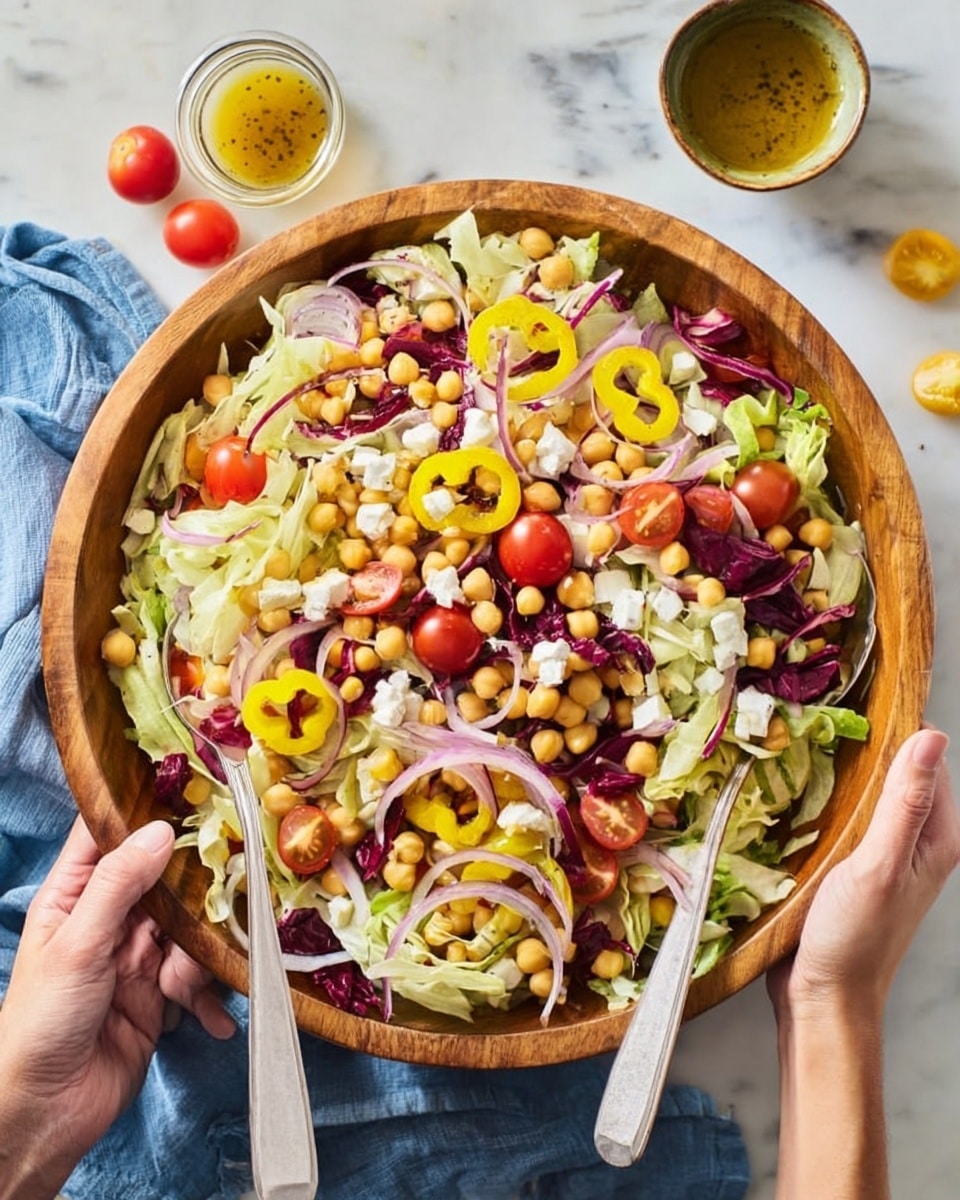 A large wooden bowl filled with a colorful salad on a white marbled surface with a blue cloth nearby, containing a vibrant mix of shredded pale green lettuce and dark purple radicchio as the base layer, topped with round yellow banana pepper rings, light beige chickpeas, small white cheese cubes, and halved bright red cherry tomatoes. Thin slices of red onion are scattered on top, adding a slight purple contrast. Two light gray salad spoons are held by a woman's hands, positioned at the edge of the bowl, ready to serve. A small glass bowl of golden dressing with a spoon sits beside the salad, and a few whole cherry tomatoes rest on the table near the bowl. photo taken with an iphone --ar 4:5 --v 7