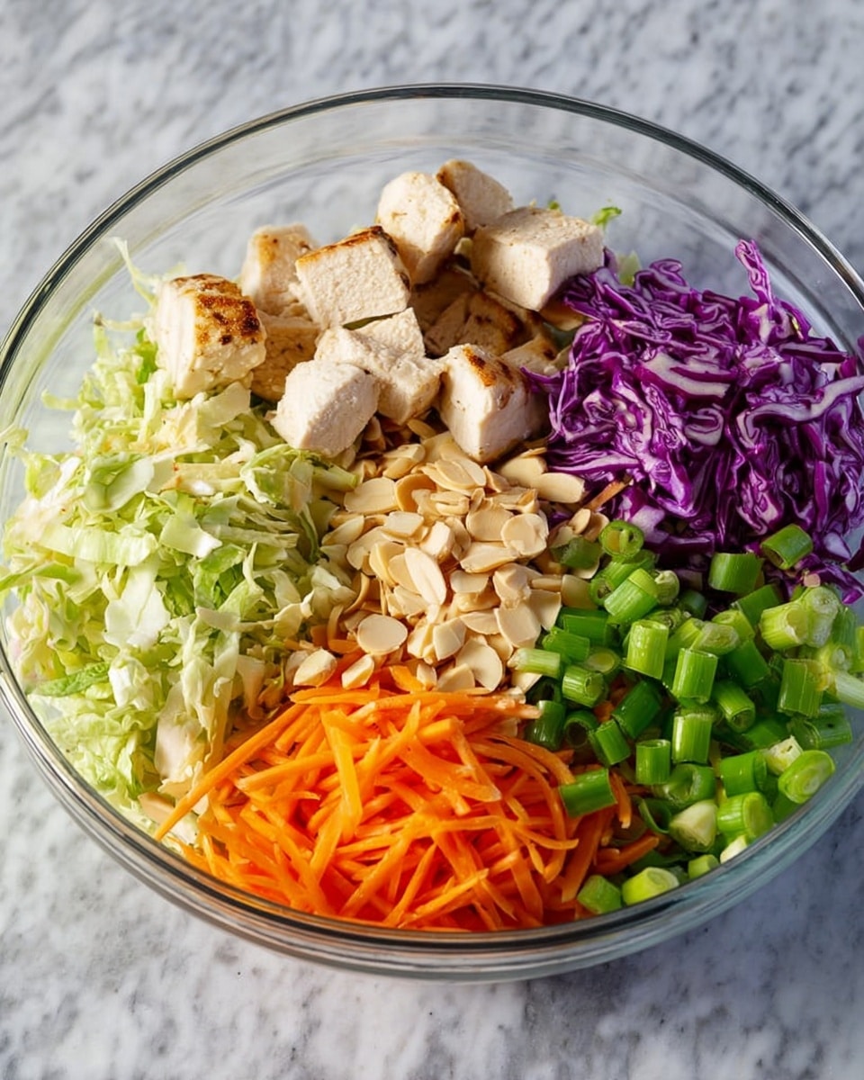 A clear bowl holds six layers of fresh salad ingredients arranged separately in neat sections. Starting from the left, there are chunks of cooked white chicken with light brown grill marks. Next to it is a bed of shredded light green cabbage, followed by finely shredded purple cabbage. To the right of the cabbage is a pile of sliced green onions. Below the chicken and cabbage sections are bright orange shredded carrots topped with slivered light beige almonds. The bowl sits on a white marbled surface. The overall look is colorful and fresh, showing the texture of each ingredient clearly. Photo taken with an iphone --ar 4:5 --v 7