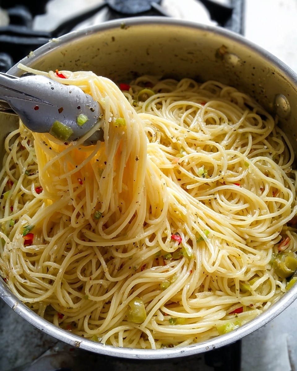 A stainless steel pot filled with cooked spaghetti that is light yellow in color with a slight shine, mixed evenly with green olive slices and small bits of red pepper scattered throughout. The surface of the noodles has visible black pepper specks for seasoning. A pair of silver kitchen tongs lifts a section of the spaghetti from the left side of the pot, showing the long, smooth strands twisting in mid-air. The pot is placed on a white marbled surface with some stove edges visible in the background. photo taken with an iphone --ar 4:5 --v 7