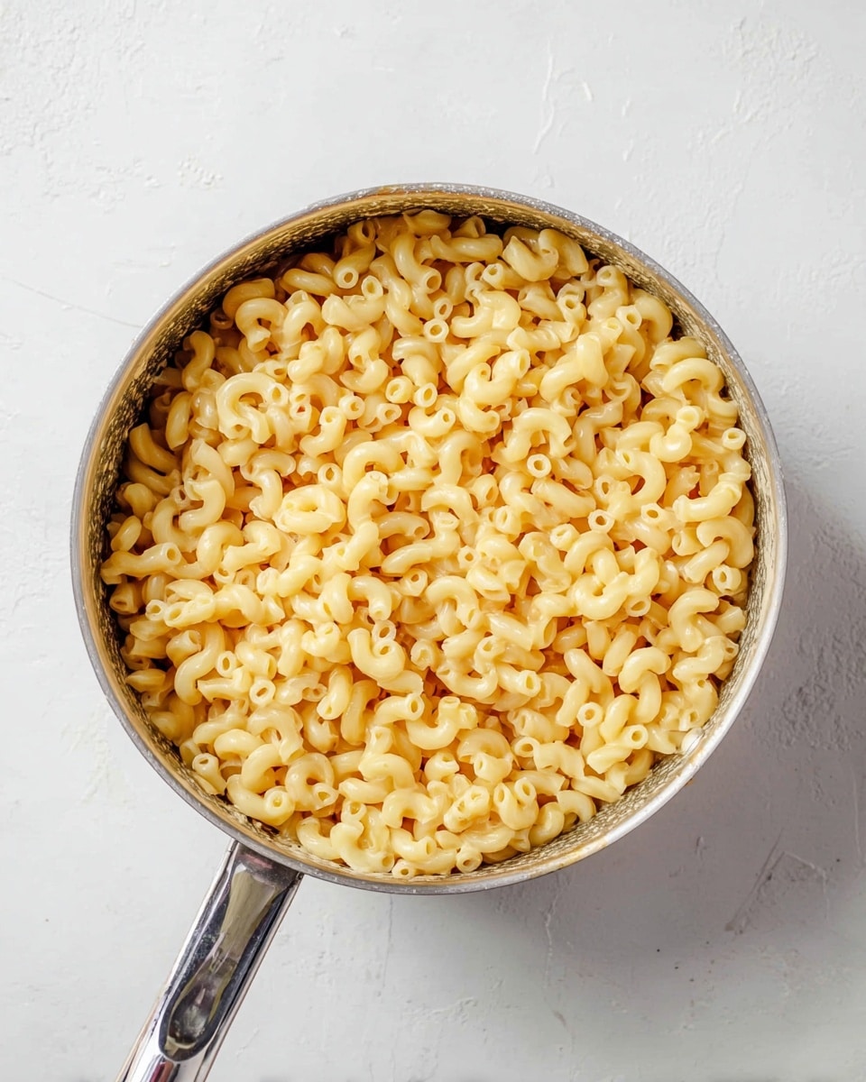 A silver saucepan filled with one layer of cooked elbow macaroni pasta, which is pale yellow and smooth in texture, sits on a white marbled surface. The macaroni pieces are tightly packed and evenly cover the bottom of the pan, with their small curved tubes showing a soft shine. The pan handle extends forward, shiny and metallic. photo taken with an iphone --ar 4:5 --v 7