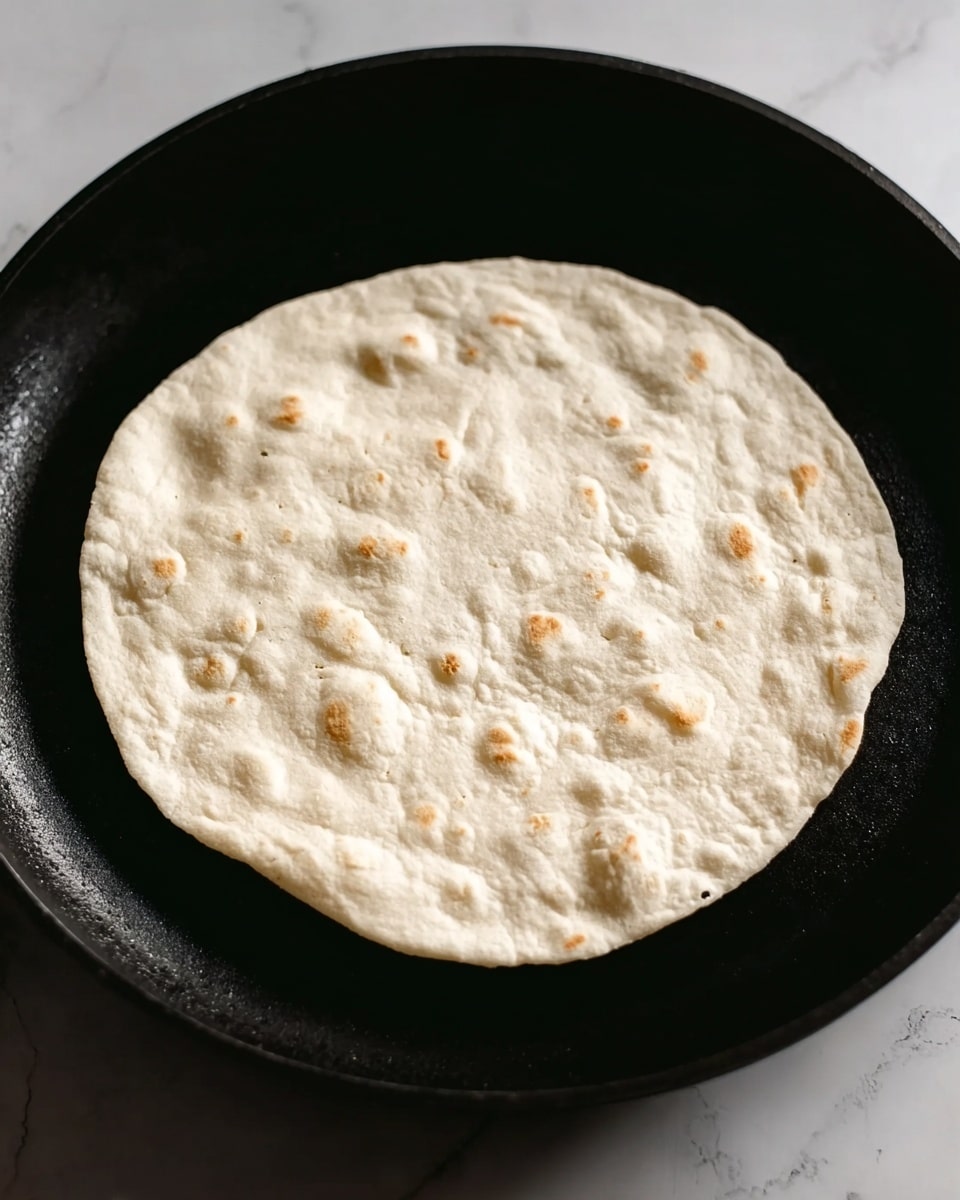 A single round flatbread with a pale, slightly bubbled surface is placed in the center of a black pan. The flatbread is light beige with some small air pockets giving it a soft texture. The black pan contrasts with the flatbread’s light color, and the background shows a white marbled surface. photo taken with an iphone --ar 4:5 --v 7
