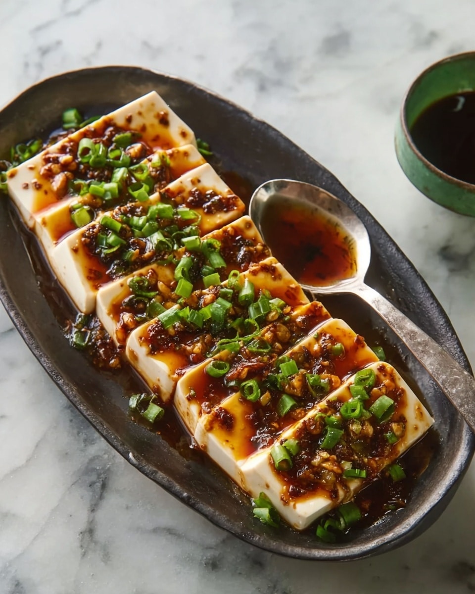 The dish is served on a dark, oval-shaped plate placed on a white marbled surface. It has eight rectangular pieces of soft tofu arranged in a single layer, each topped with a thick, glossy brown sauce. The sauce is mixed with small chopped garlic pieces and green chopped spring onions scattered evenly across the tofu. A large silver spoon lies on the right side of the plate, slightly dipped under the tofu. In the background, there is a small white bowl with a green rim containing a dark liquid. Photo taken with an iphone --ar 4:5 --v 7