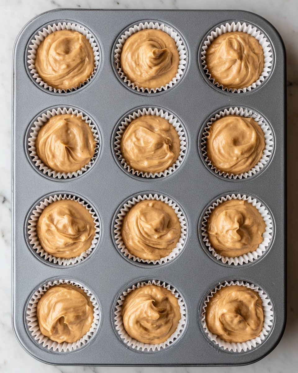 A top-down view of a gray metal muffin tray holding twelve cupcake liners filled with light brown batter. Each liner is white with ridges, and the batter is swirled unevenly, showing soft peaks and a smooth, creamy texture. The tray is placed on a white marbled surface, which contrasts with the darker tray and batter. The batter fills each liner about three-quarters full, ready to be baked. photo taken with an iphone --ar 4:5 --v 7