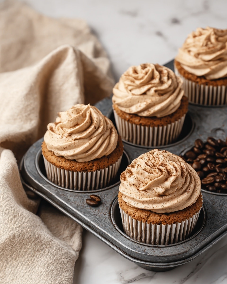 The image shows five cupcakes in a dark metal baking tray with coffee beans scattered in one empty spot. Each cupcake has a single brown cake layer topped with a thick swirl of light brown frosting sprinkled lightly with darker powder. The cupcakes are in ribbed paper liners, and the tray sits on a white marbled surface with a beige cloth draped to one side. Photo taken with an iphone --ar 4:5 --v 7