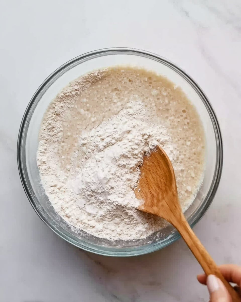 A clear glass bowl filled with a mixture of white powder and light beige liquid, creating a lumpy and foamy texture on the surface. A wooden spoon is stirring the mixture, held by a woman's hand, with some powder still visible on the surface above the liquid. The bowl is placed on a white marbled surface. photo taken with an iphone --ar 4:5 --v 7