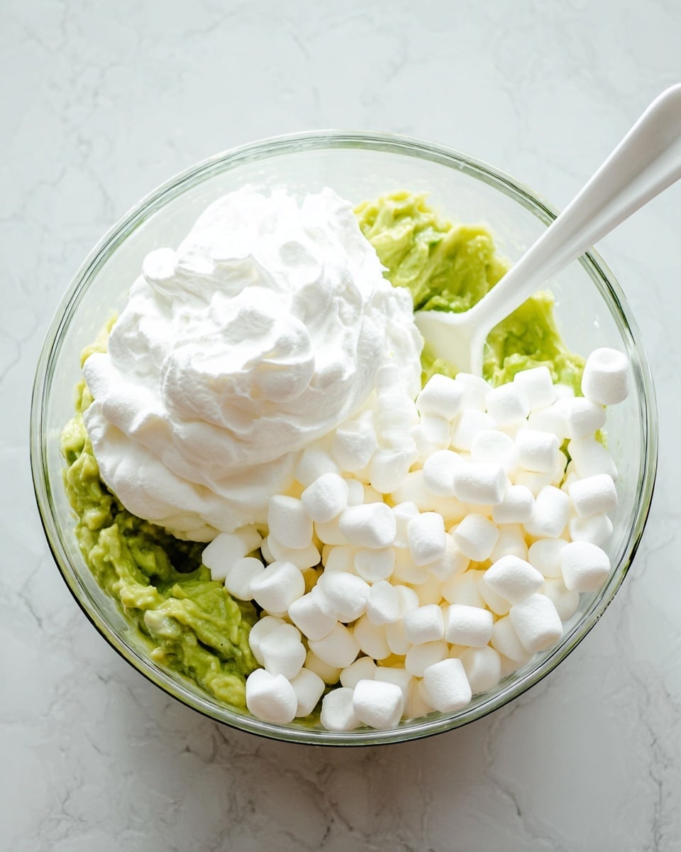 A clear glass bowl sits on a white marbled surface, filled with three distinct layers: mashed green avocado on the bottom left, a large dollop of white whipped cream on the top left, and a pile of small white marshmallows on the right side. A white spoon rests inside the bowl, leaning against the side, partially touching the avocado layer. The colors of the bowl's contents contrast clearly, with the bright green, pure white whipped cream, and soft white marshmallows all visible in the same frame. Photo taken with an iphone --ar 4:5 --v 7