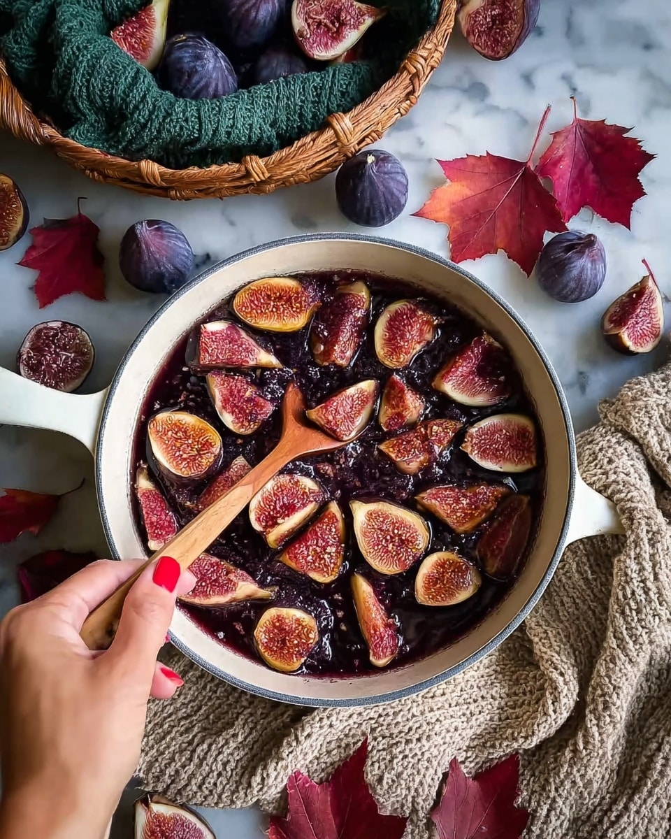 A white pan filled with layers of dark purple and red sliced figs that show their pinkish-red inner flesh. The figs are glistening, suggesting they are being cooked or softened. A woman's hand with red nail polish stirs the fruit in the pan using a wooden spoon. Around the pan, there are whole figs with dark purple skin, red autumn leaves, a green knitted cloth inside a woven basket, and a beige knitted cloth placed on a white marbled surface. Photo taken with an iphone --ar 4:5 --v 7