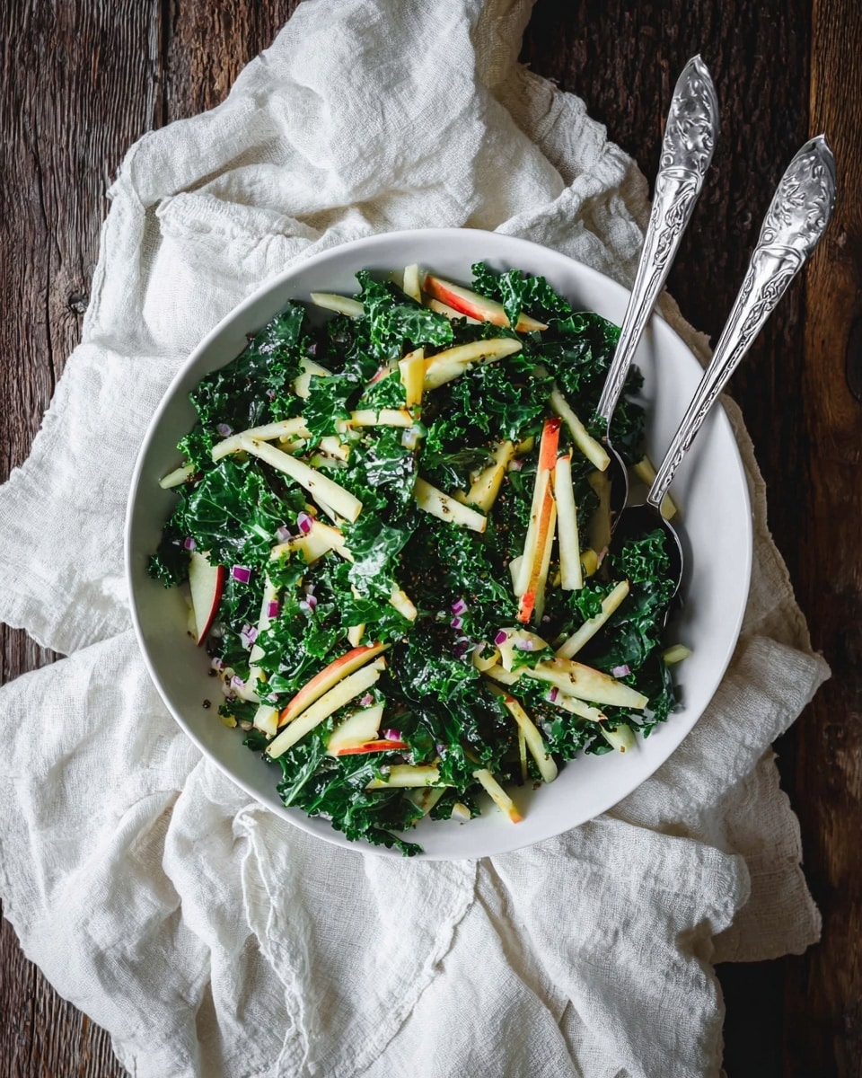 A white bowl filled with a fresh salad showing two main layers: the bottom layer of dark green curly kale leaves with a textured, leafy look, and the top layer of long, thin, pale yellow strips of apple with red edges scattered throughout. Small bits of purple onion and tiny dark poppy seeds are mixed in, adding more texture and color contrast. Two silver serving tongs with round handles rest inside the bowl, partially covered by the salad. The bowl is placed on a soft, crumpled off-white cloth, all set against a dark wooden table with visible grain patterns. Photo taken with an iphone --ar 4:5 --v 7
