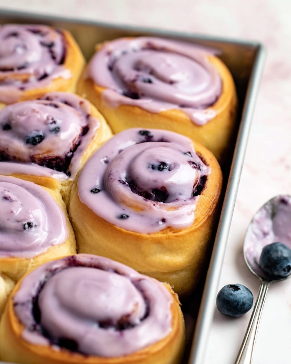 The image shows a close-up of six soft, golden-brown cinnamon rolls with a smooth, light purple glaze on top that swirls gently over each roll. The rolls are arranged in two rows in a rectangular metal pan. The glaze has small dark specks, adding texture and depth. In the background, there are three blueberries and a silver spoon with some purple glaze on a white marbled surface. The scene is bright and fresh, focused on the shiny glaze and fluffy texture of the rolls. photo taken with an iphone --ar 4:5 --v 7