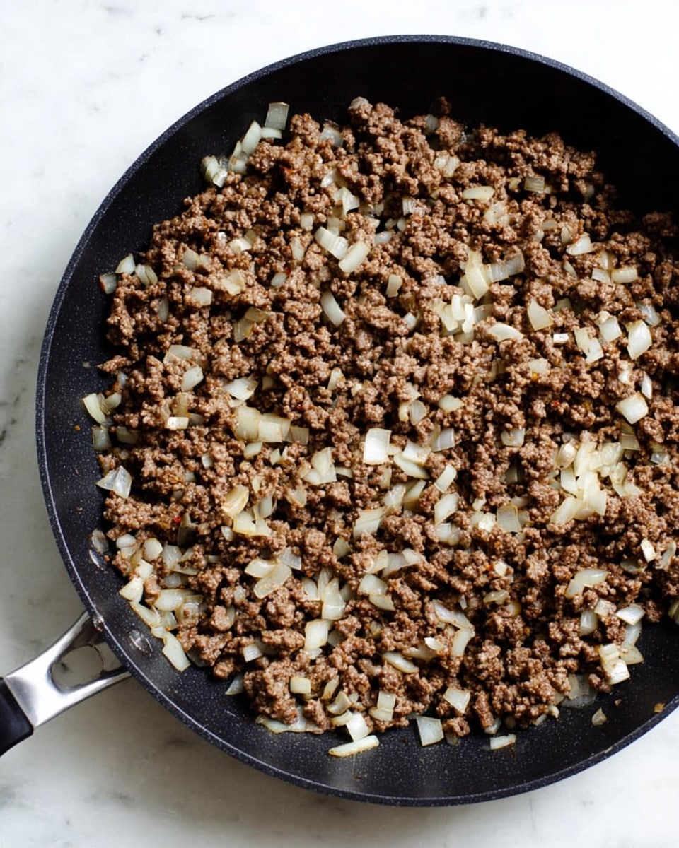 A black pan filled with cooked ground meat mixed with chopped white onions. The meat is brown with a slightly crispy texture, while the onions are translucent with soft edges, scattered evenly throughout the meat. The pan handle is visible on the left side, resting on a white marbled surface. photo taken with an iphone --ar 4:5 --v 7