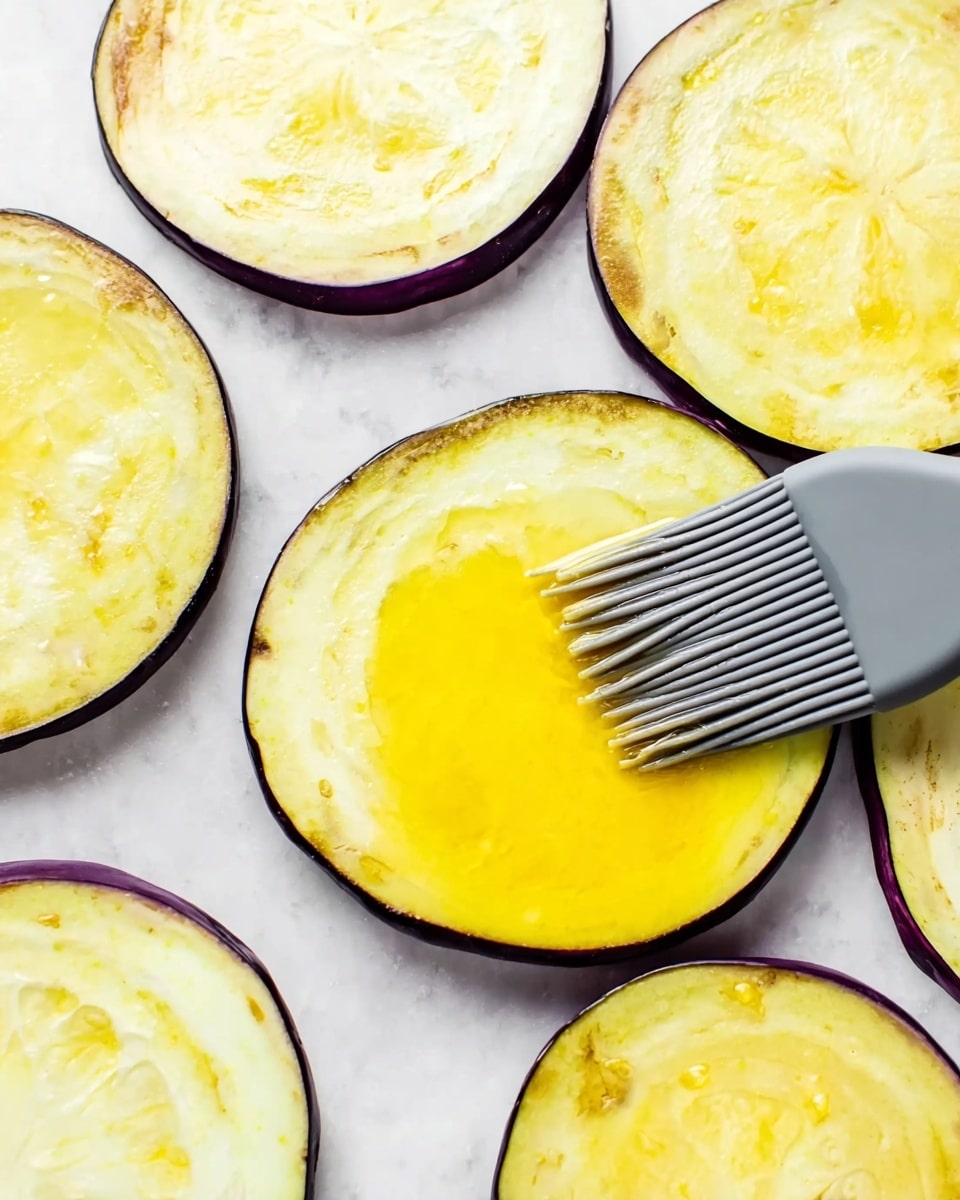 Several round slices of eggplant are laid out on a white marbled surface. Each slice has a pale yellow inside with some lighter and darker patches and a thin dark purple peel around the edges. A grey silicone brush is spreading bright yellow oil or butter over one eggplant slice in the center. The texture of the brush bristles is clear, and the shine on the yellow spread is glossy. The eggplant slices are arranged so that some slightly overlap the others. Photo taken with an iphone --ar 4:5 --v 7
