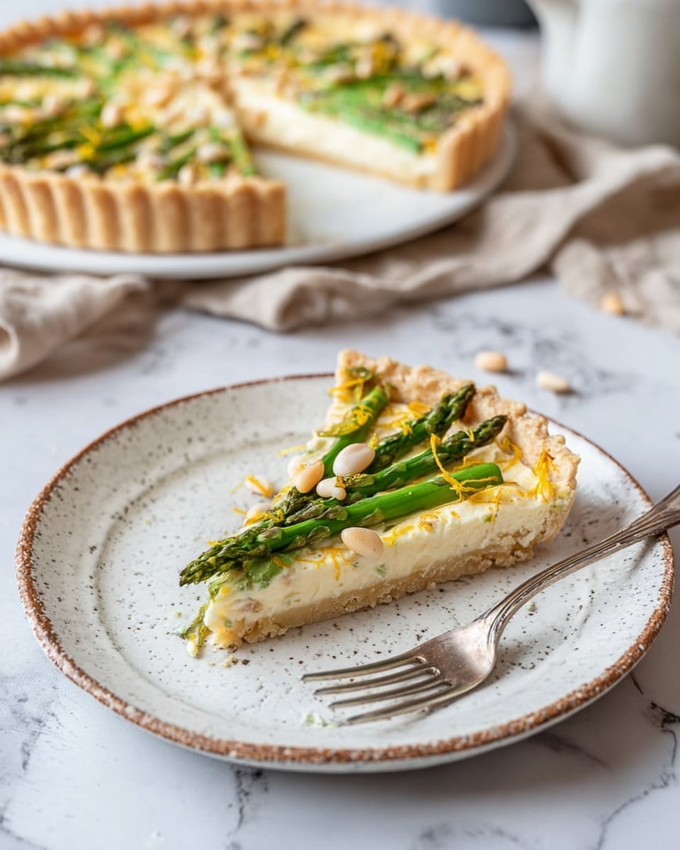 A slice of tart is shown on a white rustic plate, with a golden, flaky crust forming the base layer. On top, a creamy, pale cheese layer covers the crust evenly, topped with bright green asparagus spears placed in a neat row. Small, light tan pine nuts are scattered over the asparagus, with tiny bits of yellow zest sprinkled on top for color. A silver fork lies next to the tart slice on the plate. The background features the rest of the tart on a white marbled surface, blurred softly, with a beige cloth nearby. Photo taken with an iphone --ar 4:5 --v 7
