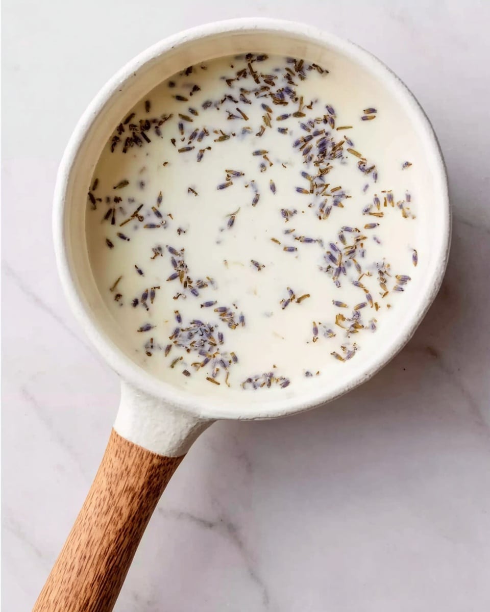A top-down view of a white pot with a wooden handle resting on a white marbled surface. The pot is filled with a creamy white liquid, sprinkled evenly with small dried lavender petals scattered throughout. The texture of the liquid is smooth and contrasts with the delicate lavender pieces. The handle extends diagonally out from the pot, adding a natural wood tone to the image. Photo taken with an iphone --ar 4:5 --v 7