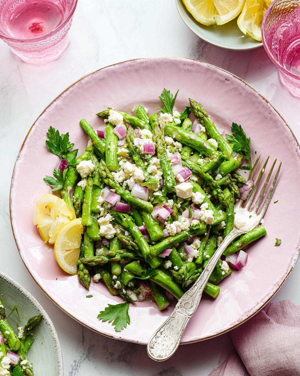 The image shows a pink plate filled with a fresh asparagus salad consisting of three main layers. The bottom layer is made of bright green asparagus pieces, arranged loosely across the plate. On top of the asparagus, there is a layer of chopped red onions in small square pieces, scattered unevenly. The top layer has small white crumbles of cheese sprinkled all over, with two thin lemon slices placed near the edge on one side, and green parsley leaves placed as garnish across the salad. A silver fork rests on the right side of the plate, partially on the salad. The plate is set on a white marbled surface, and in the background, there is a clear glass with pink liquid and another bowl with lemon slices visible. Photo taken with an iphone --ar 4:5 --v 7