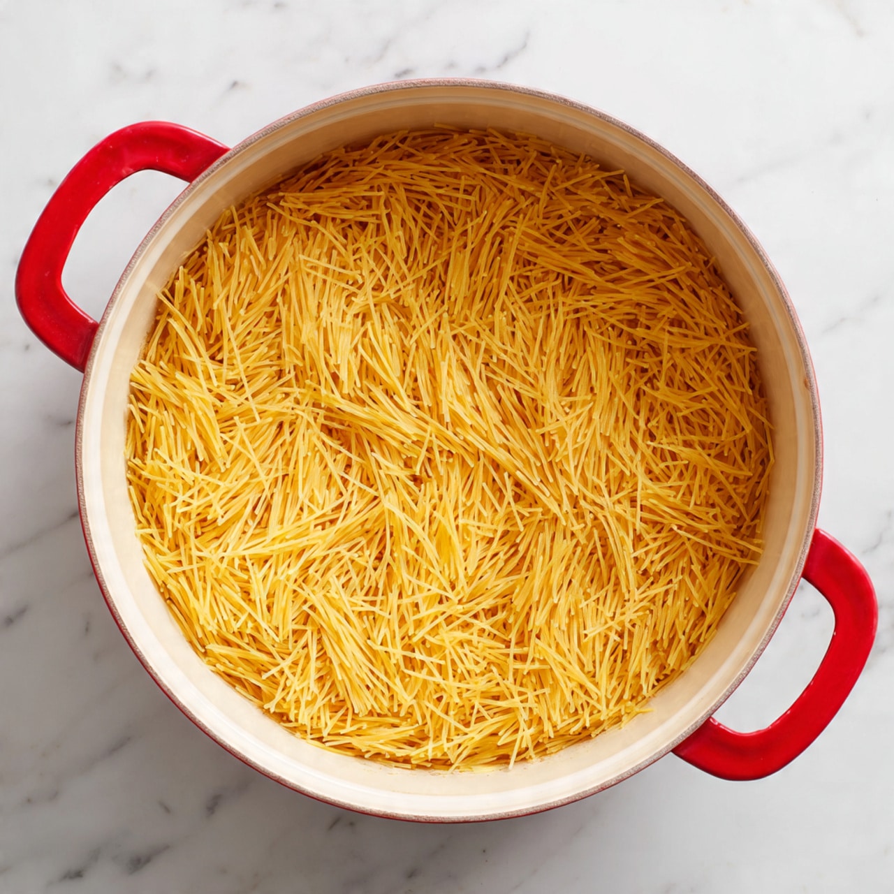 A round white pot with bright red handles sits on a white marbled surface. Inside the pot is one even layer of thin, short, dry yellow pasta strands spread thinly across the bottom, showing a few slightly browned bits on the edges. The pasta has a dry, firm texture and fills the base of the pot without overlapping. Photo taken with an iphone --ar 4:5 --v 7