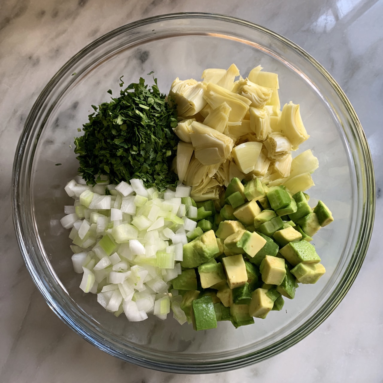 A clear glass bowl sits on a white marbled surface, filled with five separate piles of chopped ingredients that are not mixed. In the top right, there are chunks of pale yellow artichoke hearts with a soft texture. At the bottom right, diced green avocado pieces with smooth, creamy texture. At the bottom center, finely chopped white onion pieces, small and slightly translucent. At the bottom left, light green diced celery with a crisp appearance. At the top left, a small pile of finely chopped fresh dark green herbs. Photo taken with an iphone --ar 4:5 --v 7