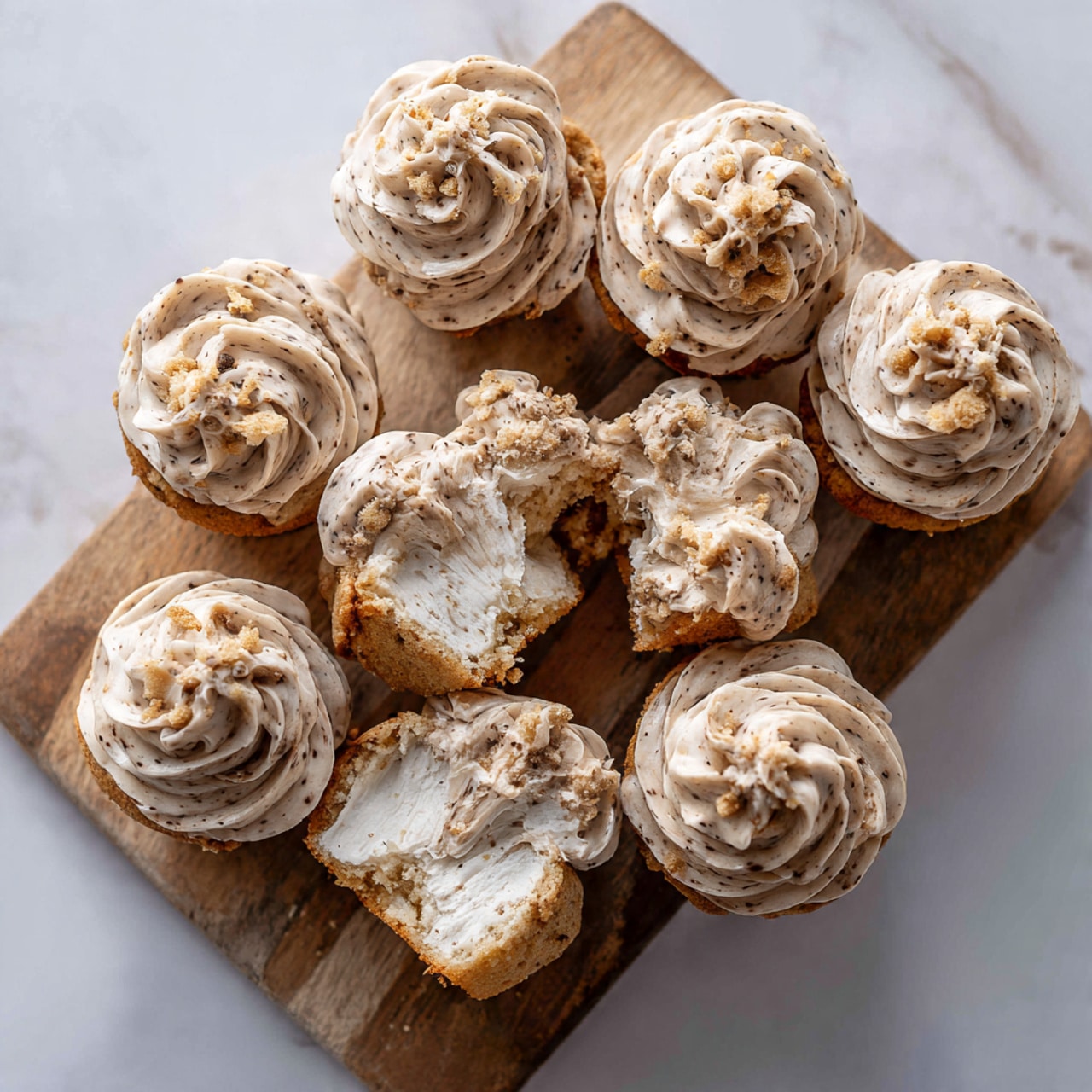 The image shows a wooden board on a white marbled surface holding eleven small desserts that look like muffins topped with a thick, swirled, light brown frosting with specks throughout. One of the desserts is broken in half, revealing a soft, fluffy white inside with a golden brown base and some crumbs around. The desserts are arranged close together, and the board has a rustic, slightly rough texture with natural wood grain visible. The photo was taken with an iphone --ar 4:5 --v 7