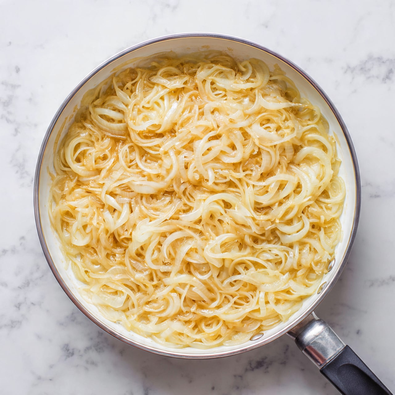 A round pan filled with a thick layer of cooked thinly sliced onions, pale yellow and shiny with a soft texture. The onions are spread evenly inside the pan, with their delicate curved shapes creating a subtle pattern. The pan has a white interior with a silver metal edge and handle, and a black handle extending out. The pan rests on a white marbled surface, adding a clean and bright background to the image. Photo taken with an iphone --ar 4:5 --v 7