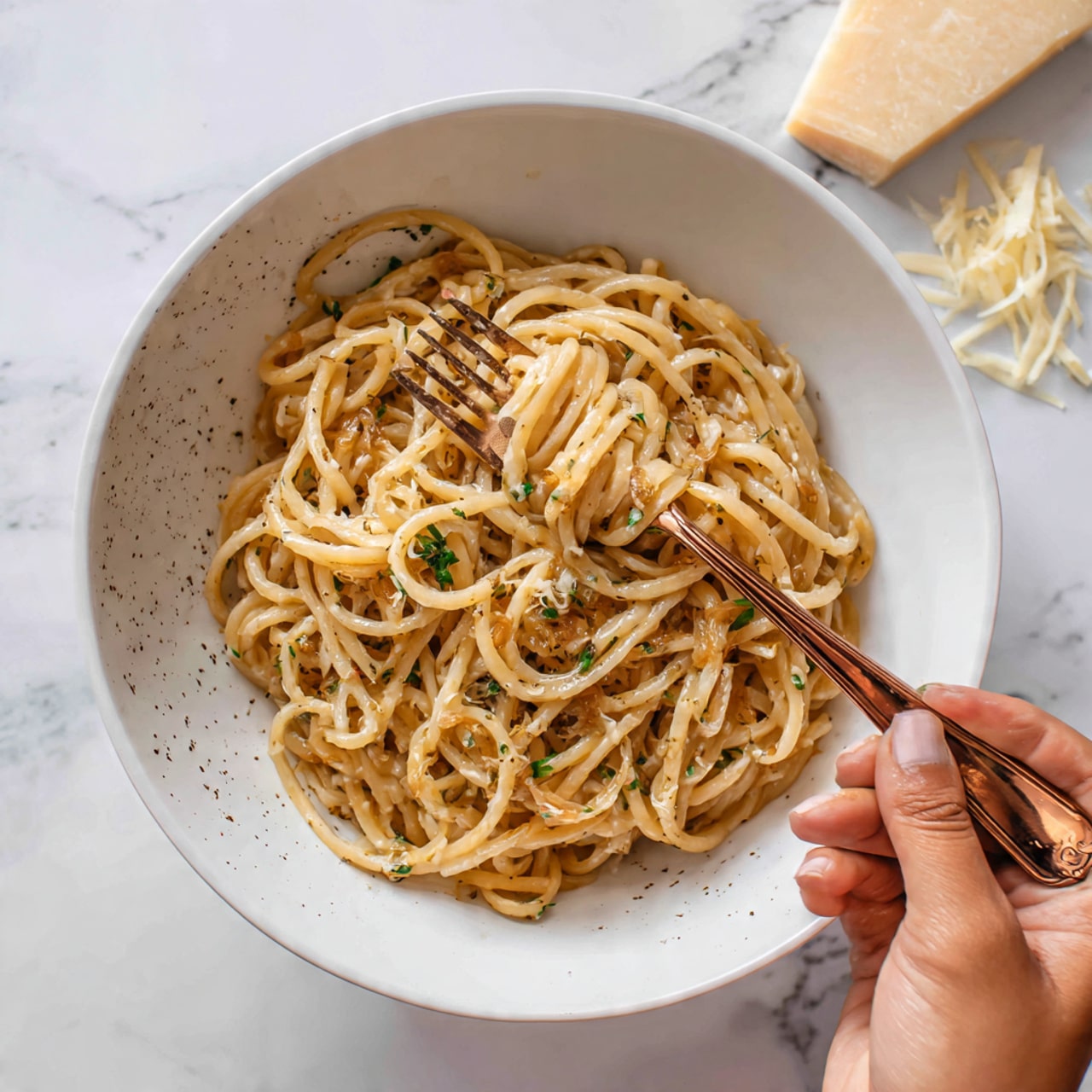 A close-up of thick, pale yellow noodles that are soft and slightly twisted, covered in a light brown sauce with small bits of herbs sprinkled on top, served in a large white bowl with small dark specks. A woman’s hand holds a copper fork near the center of the image, with a wrapped bundle of noodles tightly twirled around the fork’s tines. The background features a white marbled texture with a few shreds of pale cheese resting nearby. Photo taken with an iphone --ar 4:5 --v 7