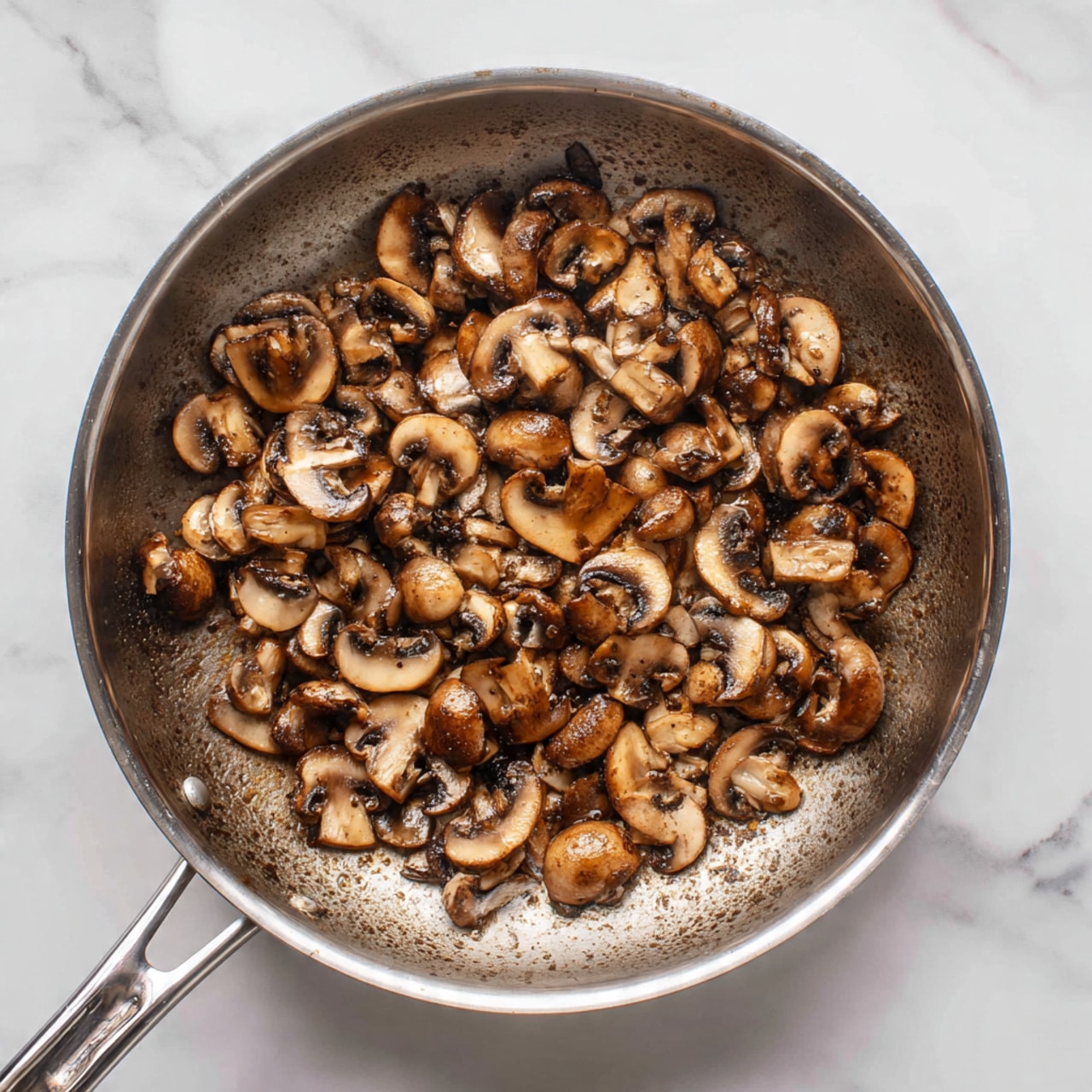 A top view of a stainless steel pan with many small brown cooked mushrooms spread inside. The mushrooms have a slightly shiny texture and are unevenly placed so some of the pan bottom is visible. The pan sits on a white marbled surface, showing subtle grey veins and a clean, bright look. The handle of the pan is visible on the left side. photo taken with an iphone --ar 4:5 --v 7
