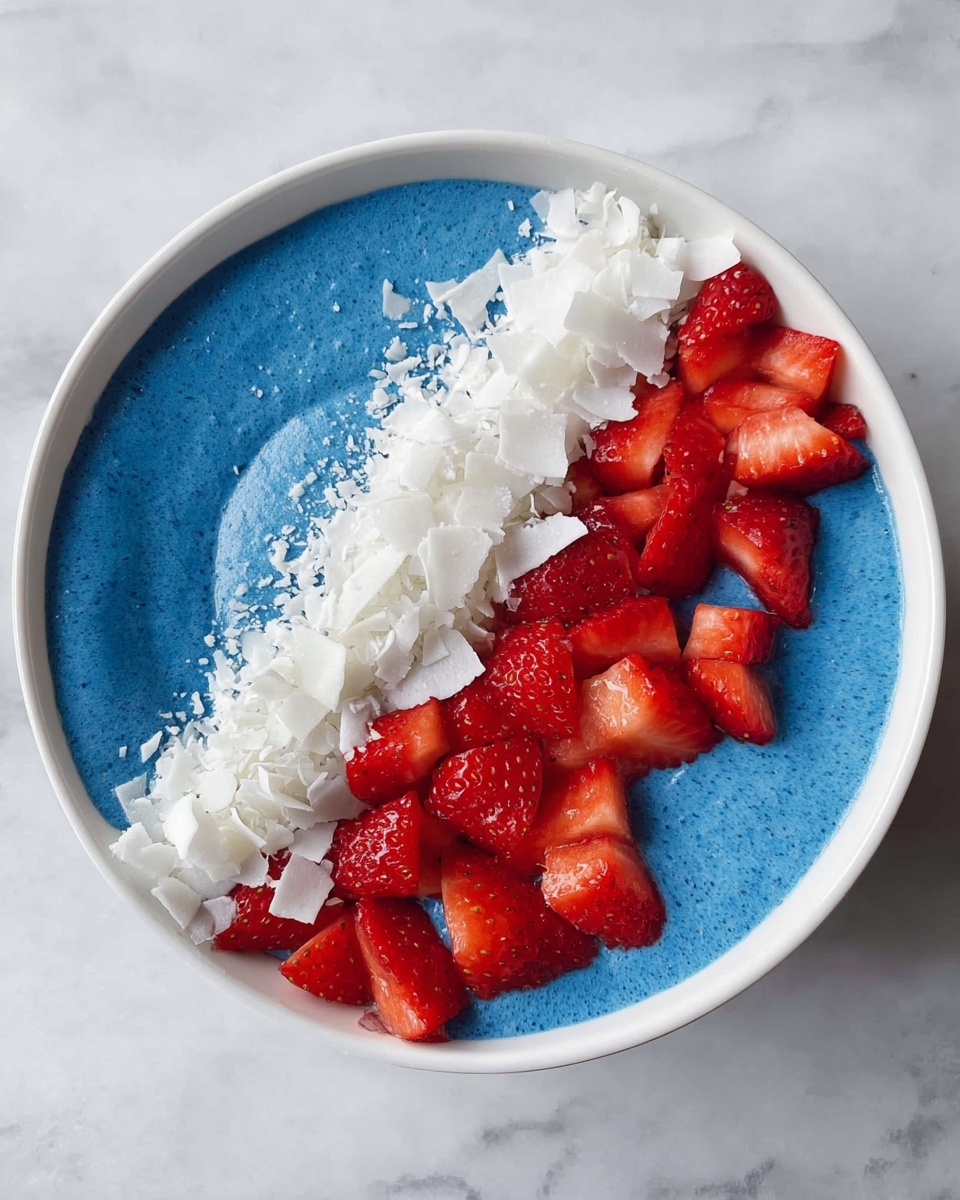 A white bowl filled with a smooth, thick blue base covers about two-thirds of the bowl, while the left third is topped with two layers: bright red chopped strawberries forming one layer on the left edge, and a strip of white coconut flakes next to the strawberries, covering part of the blue base. The bowl is placed on a white marbled surface. photo taken with an iphone --ar 4:5 --v 7