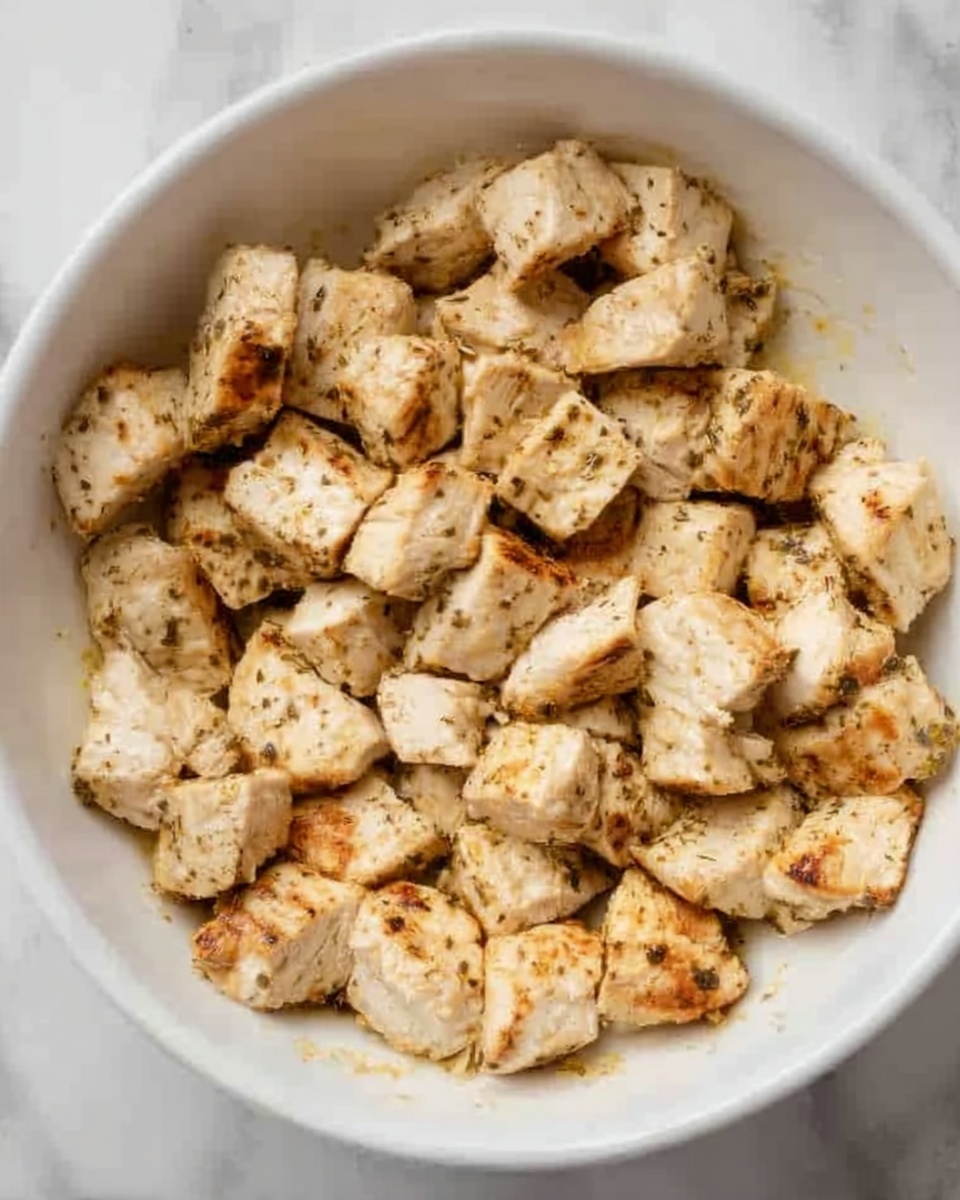 The image shows a close-up of small, browned cubes of chicken cooked in a white bowl. The chicken pieces are lightly browned with some char marks and are spread evenly inside the bowl. The chicken looks juicy and soft with a simple seasoning showing small specks of herbs or spices. The bowl is placed on a white marbled surface. photo taken with an iphone --ar 4:5 --v 7