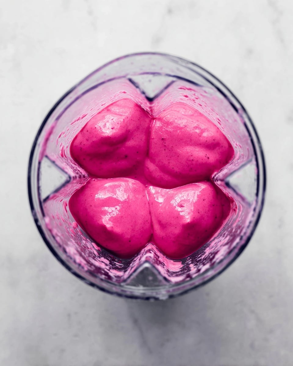 A clear blender container is shown from above, filled nearly to the top with a bright pink thick smoothie mixture that is smooth and shiny. The mixture forms four rounded lobes with a slightly uneven surface and some small darker specks inside. The blender is placed on a white marbled surface with soft natural light highlighting the glossy texture of the smoothie photo taken with an iphone --ar 4:5 --v 7