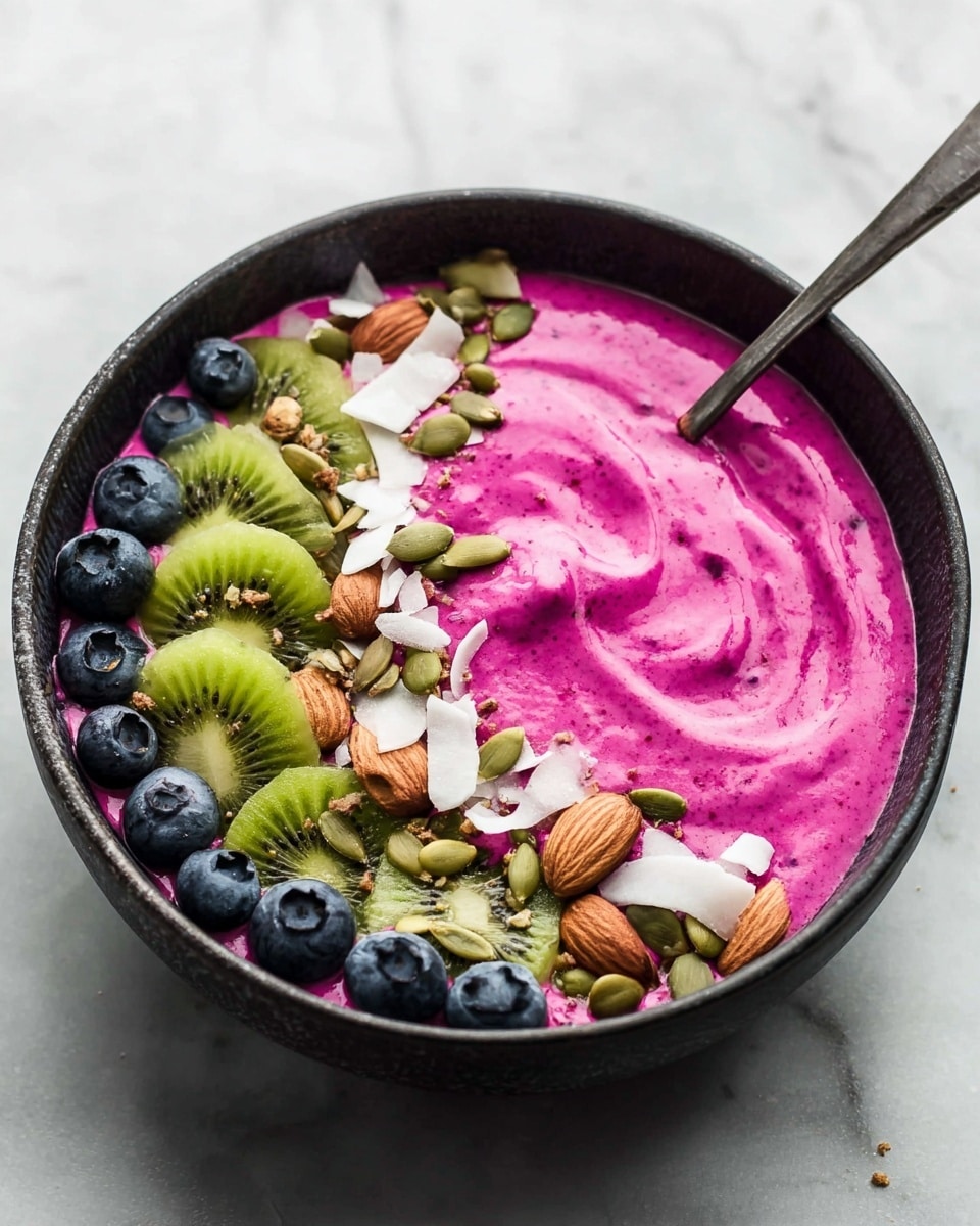 A dark bowl filled with a bright pink smoothie layer, smooth with swirl textures on the surface. On top, there are layers of toppings arranged in curved lines: green pumpkin seeds, whole brown almonds, thin white coconut flakes, and round green kiwi slices with black seeds in the center. Scattered blueberries add dark blue pops of color around the edges. The bowl sits on a white marbled texture surface. A metal spoon rests inside the bowl near the top edge. Photo taken with an iphone --ar 4:5 --v 7