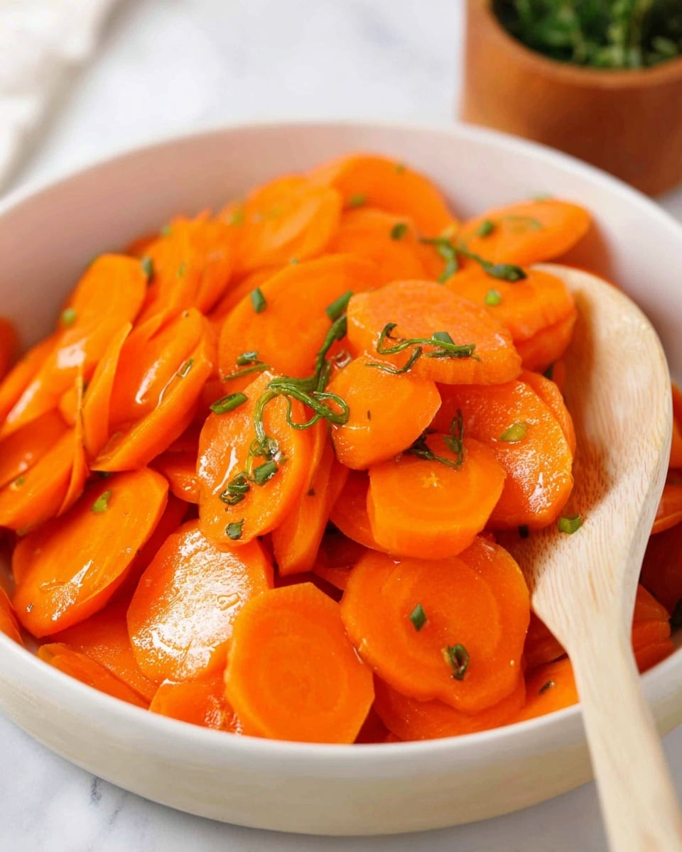 The image shows a white bowl filled with bright orange carrot slices, evenly cut and cooked to a soft texture with a slight shine. The carrot slices are mixed with small green chive pieces sprinkled on top for color contrast. A light wooden spoon rests inside the bowl on the right side, touching the carrot slices. The background features a white marbled surface with a small, blurred orange pot of green herbs in the upper right corner. The overall colors are warm and fresh, focusing on the vibrant orange and green hues. photo taken with an iphone --ar 4:5 --v 7